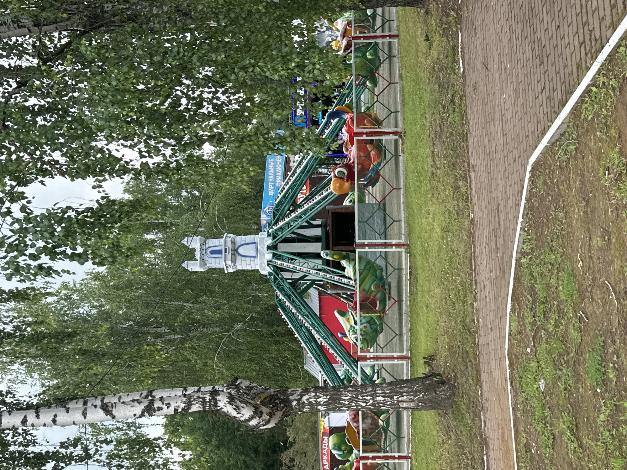 This image shows a colorful amusement park ride in Saransk, Russia. The ride features a central tower resembling a castle and several attached cars designed as fantastical creatures, such as a green dragon-like figure and a red and blue parrot-like figure. The ride is suspended on a green metal structure that rotates around the central tower. A red and green fence encloses the ride area, with a brick pathway visible in the foreground. Lush green trees and foliage surround the park, indicating an outdoor setting. Signs in Russian are visible, including one that reads "ВИРТУАЛЬНЫЕ ПРИКЛЮЧЕНИЯ" (Virtual Adventures). The overall scene suggests a playful and family-friendly atmosphere within the amusement park.
