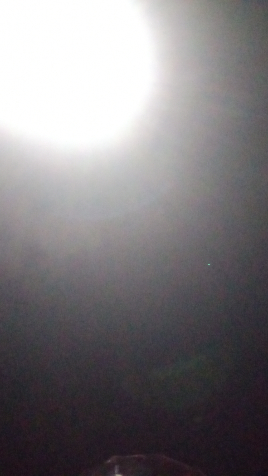 The image is a low-light, overexposed photograph that appears to be taken at night. A bright, diffused light source dominates the upper left quadrant of the image, creating a strong lens flare and washing out much of the scene. The rest of the image is dark and indistinct, with some hazy, indistinct shapes and a small, faint green dot visible in the mid-right area. The overall impression is of a blurred, abstract image, likely taken with a mobile phone camera under poor lighting conditions. Without additional context or clearer details, it is impossible to determine the specific location in Kuta, Indonesia, or any specific subjects, objects, or actions within the scene.