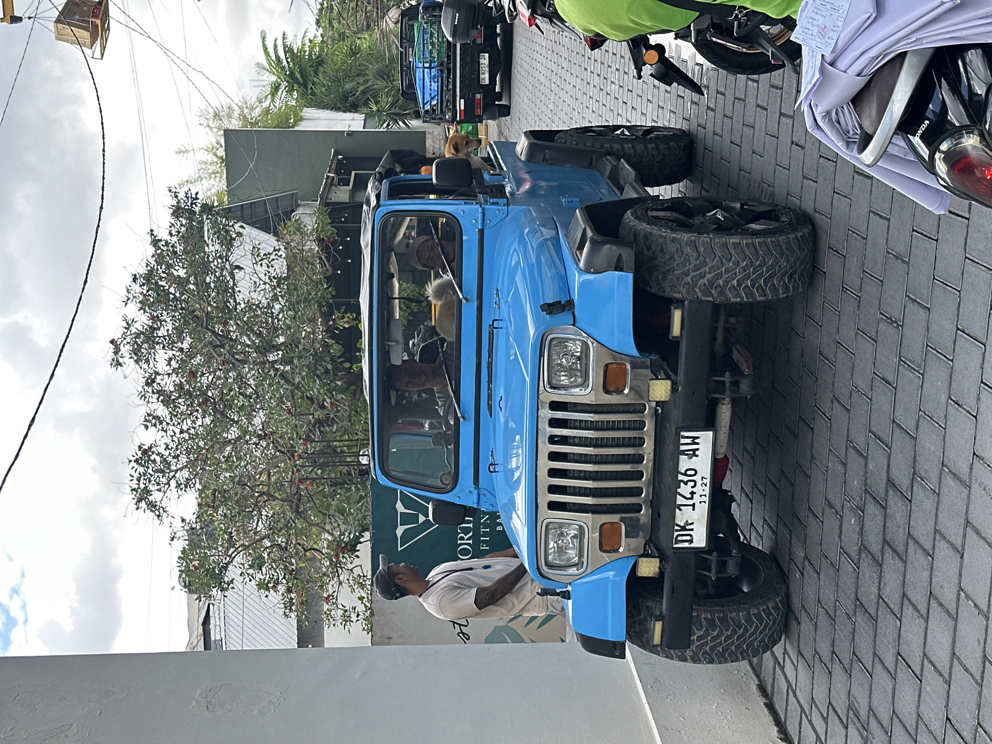 A bright blue Jeep Wrangler is parked on a cobblestone street in North Kuta, Indonesia. A man in a light-colored t-shirt and a baseball cap stands to the left of the Jeep, looking towards it. Inside the Jeep, three people are visible, two in the front and one in the back. Two dogs are also in the Jeep; one white fluffy dog sits in the passenger seat, and a tan dog with its head out the rear window. The Jeep has a license plate that reads "DK 1436 AW". In the background, a dark-colored truck with a cargo bed is parked, and behind it, a building with a green facade and trees can be seen. The sky is overcast with clouds.