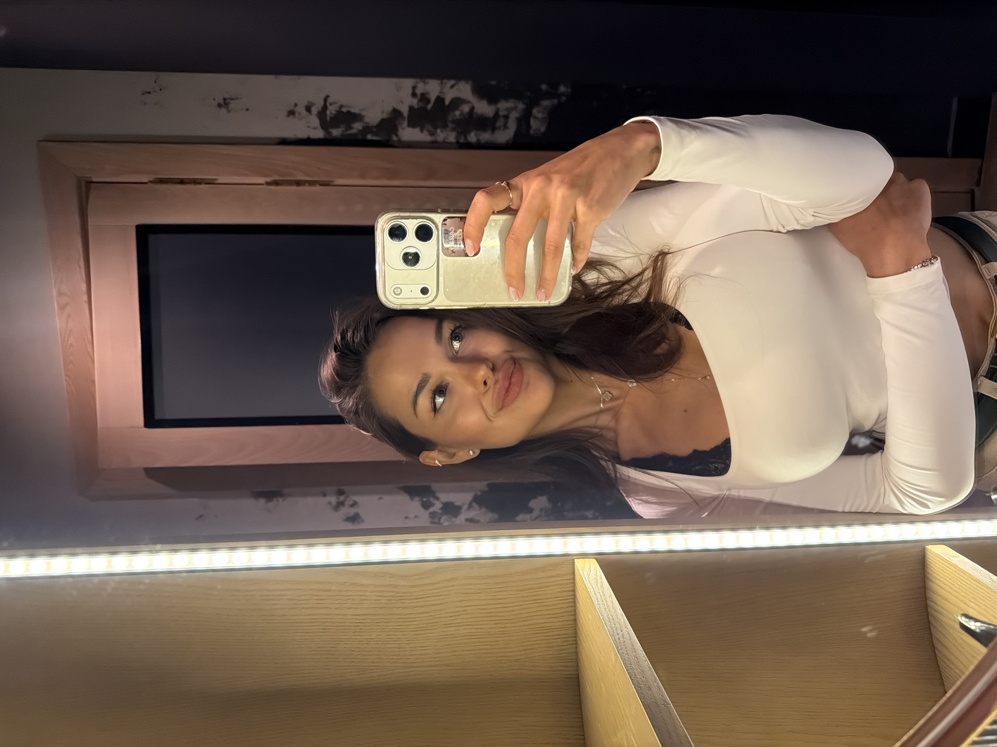A woman is taking a mirror selfie in what appears to be a dressing room or a hallway with wooden shelving and a door. She is wearing a white long-sleeved top over a black lace bra. She has long, dark hair and is looking to the side with her lips slightly pursed. She is holding a smartphone in her right hand and wearing a ring on her right index finger. The environment is dimly lit with a strip of LED lights illuminating the side of the shelving unit. The background includes a wooden door frame and dark walls with a textured finish. There is no visible indication of the location being in Saint Petersburg, Russia.