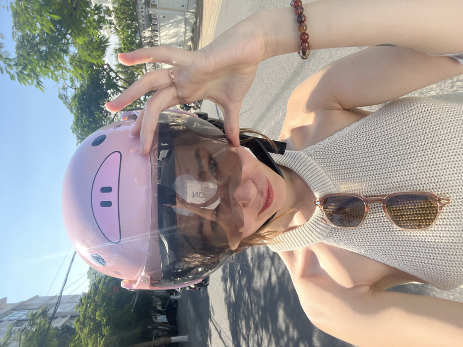 A person wearing a pink helmet with a pig face design is posing for a selfie. A hand with a ring rests near the helmet's visor. Sunglasses are draped on a beige sweater. The background features trees, buildings, and a bright blue sky. The image appears to be taken outdoors on a sunny day.