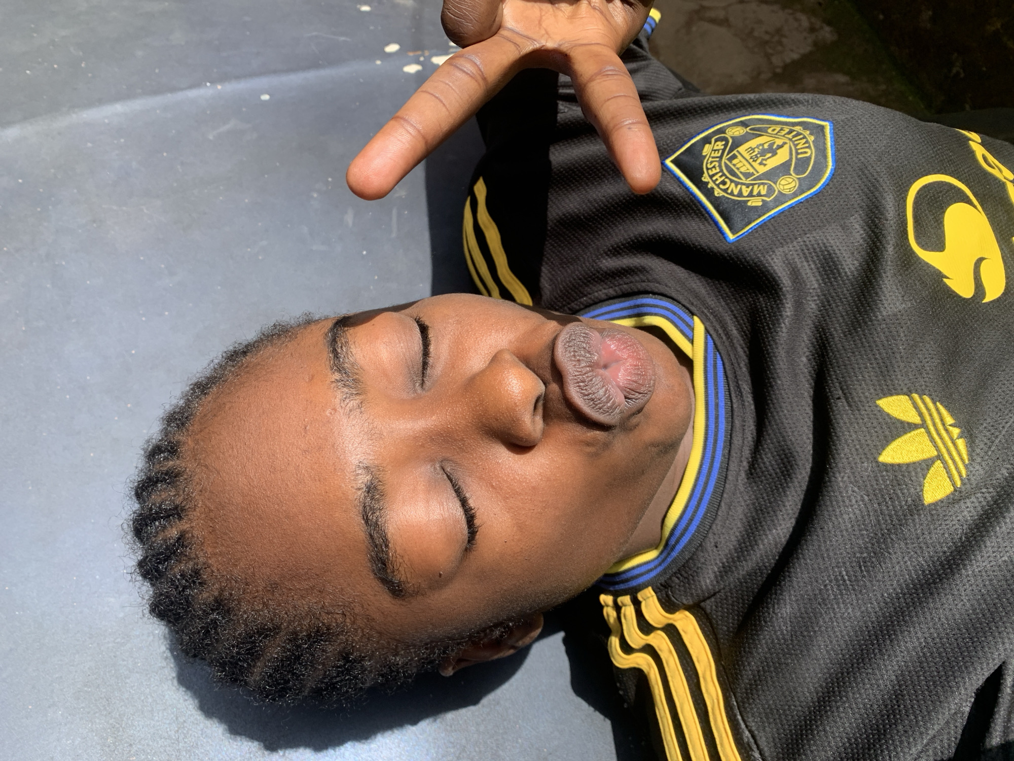 The image shows a close-up of a young Black person lying down outdoors, with their eyes closed and lips pursed. They are wearing a black jersey with yellow and blue stripes on the collar and sleeves, featuring the Manchester United logo and an Adidas logo. The person is holding up two fingers in a peace sign. The background appears to be a gray, textured surface, possibly a wall or pavement, with a shadow cast on the left side of the image. The lighting suggests it is daytime. There are no identifiable landmarks indicating a specific location in Rafin Par, Nigeria.