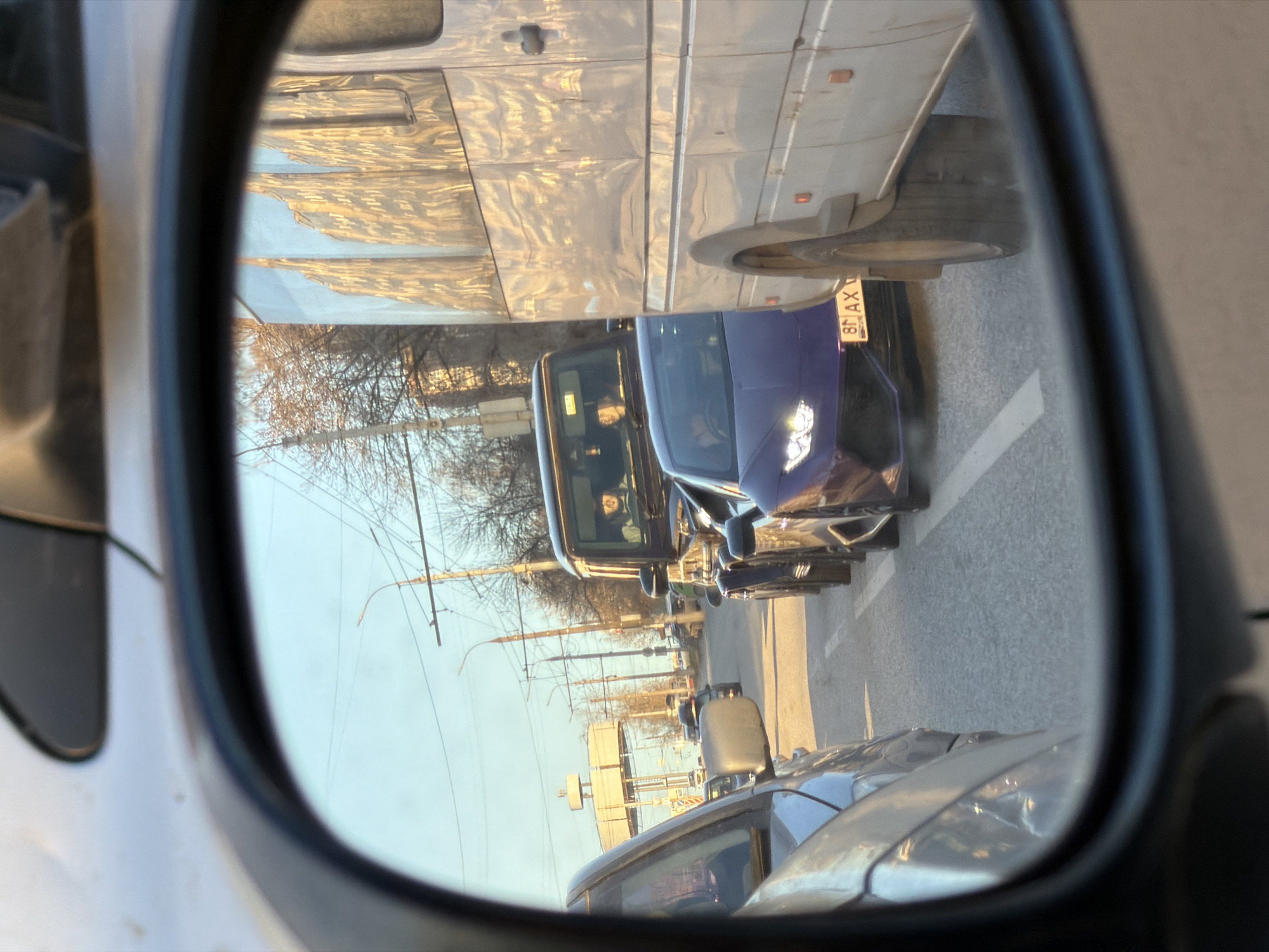 The image depicts a view from a car's side mirror. In the reflection, a street scene is visible. Several vehicles are present, including a purple car with license plate "81 AX" and a white bus. The license plate suggests the location may be in Izhevsk, Russia. Other cars are seen further down the road. Utility poles with wires and bare trees line the street. The sky is clear and light blue. A person is visible in the purple car.