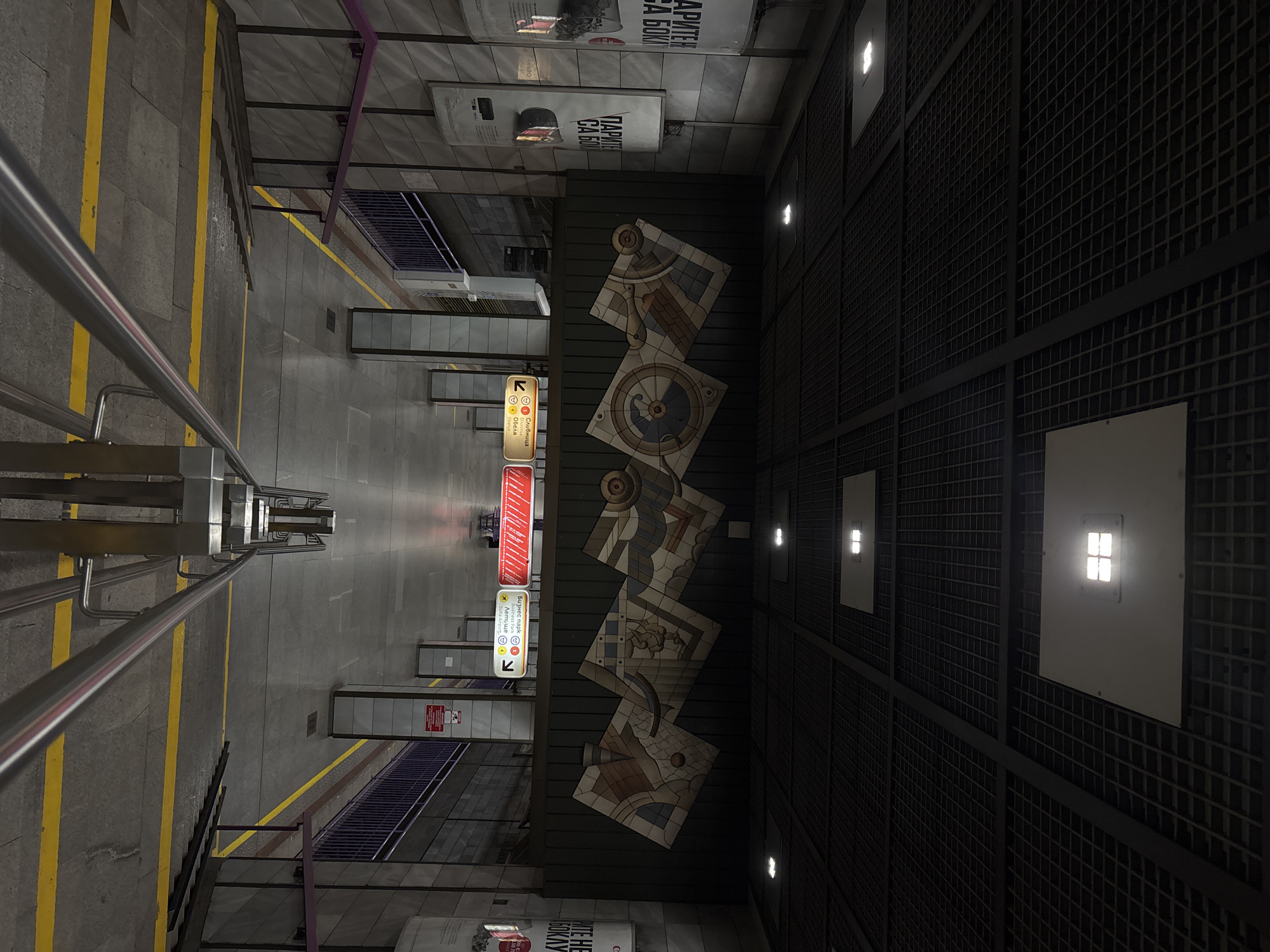 The image shows the interior of a metro station, likely in Sofia, Bulgaria. The viewer seems to be looking down the stairs from an elevated position. On the left, there are stairs with a metal handrail leading down to the platform level. Yellow lines are painted on the floor, indicating the edge of the platform and safety zones. There are signs in Bulgarian and English indicating directions to "Obelya," "Business Park," and "Sofia Airport." Along the wall on the right, a series of decorative tile panels are visible. The ceiling is a dark grid structure with embedded lights. Advertisements are posted above the tile panels. The lighting is a mix of natural and artificial, with a slightly dim and industrial feel. The scene is devoid of people.