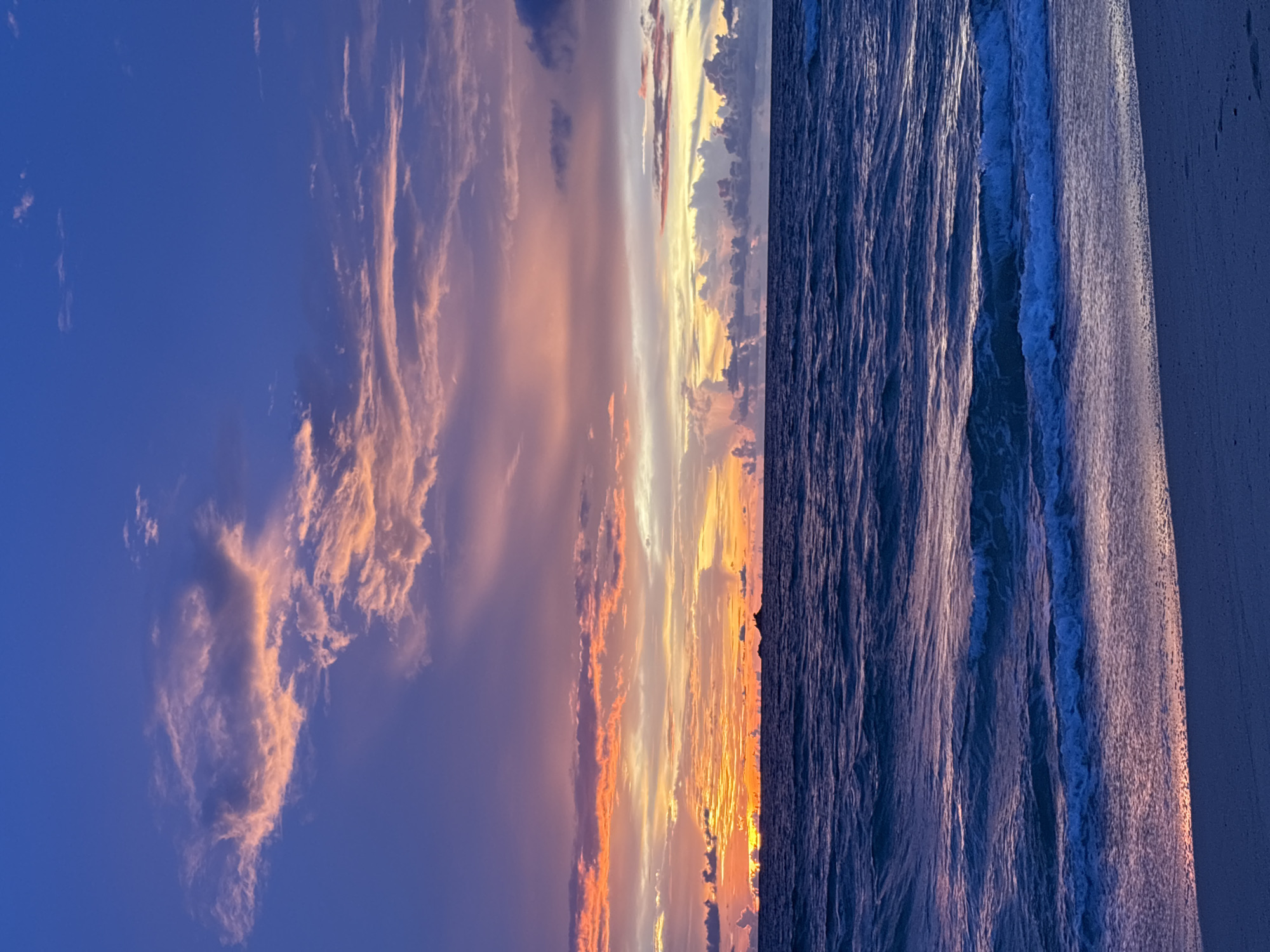 A serene sunset paints the sky with vibrant hues of purple, pink, and orange above a dark blue ocean. Gentle waves lap at a sandy beach, casting a purple reflection on the wet sand. In the distance, a small island is silhouetted against the colorful horizon. The scene is devoid of any people or man-made structures beyond the distant island. The image appears to be taken on a beach in Choeng Thale, Thailand, likely during twilight.