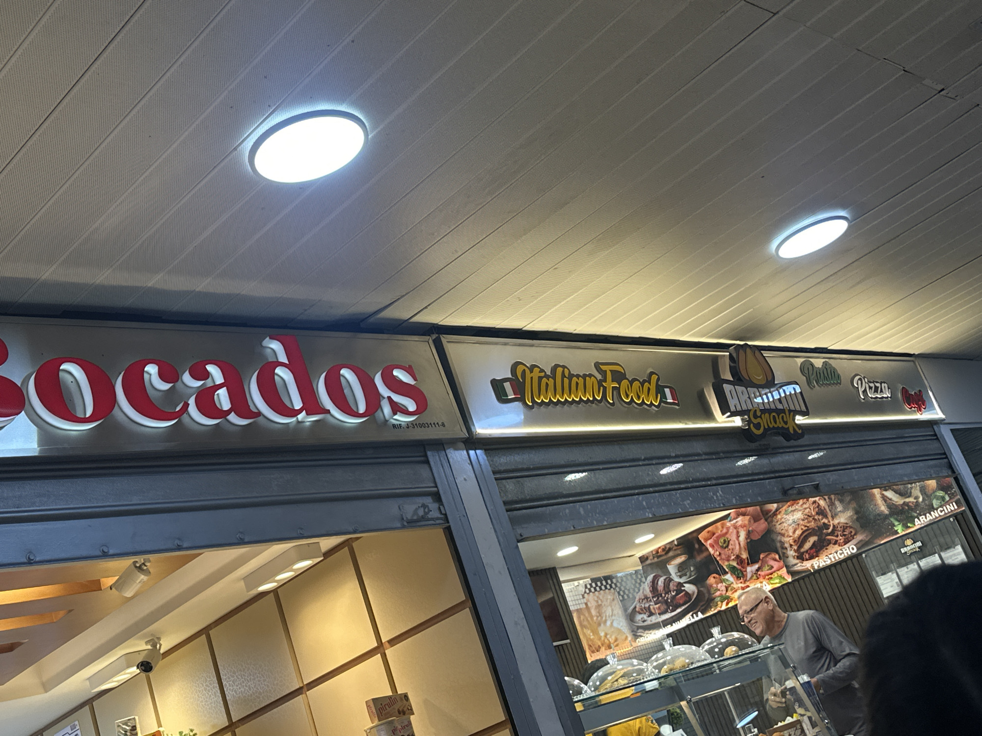 The image shows the storefront of "Bocados Italian Food Arancini Snack Pasta Pizza Cafe" in Catia La Mar, Venezuela. The red "Bocados" sign is prominent on the left. Adjacent to it is a sign with "Italian Food" written in cursive, accompanied by the flags of Italy. To the right, there is a sign reading "Arancini Snack" and another displaying "Pasta Pizza Cafe". A horizontal strip featuring images of prepared food like pasta and pizza is visible beneath the signs. An older man wearing glasses and a gray shirt is standing inside near a display case containing various foods. The shop's interior is visible through the window, revealing tiled walls. The scene is illuminated by ceiling lights. The storefront is under an overhang with paneling.