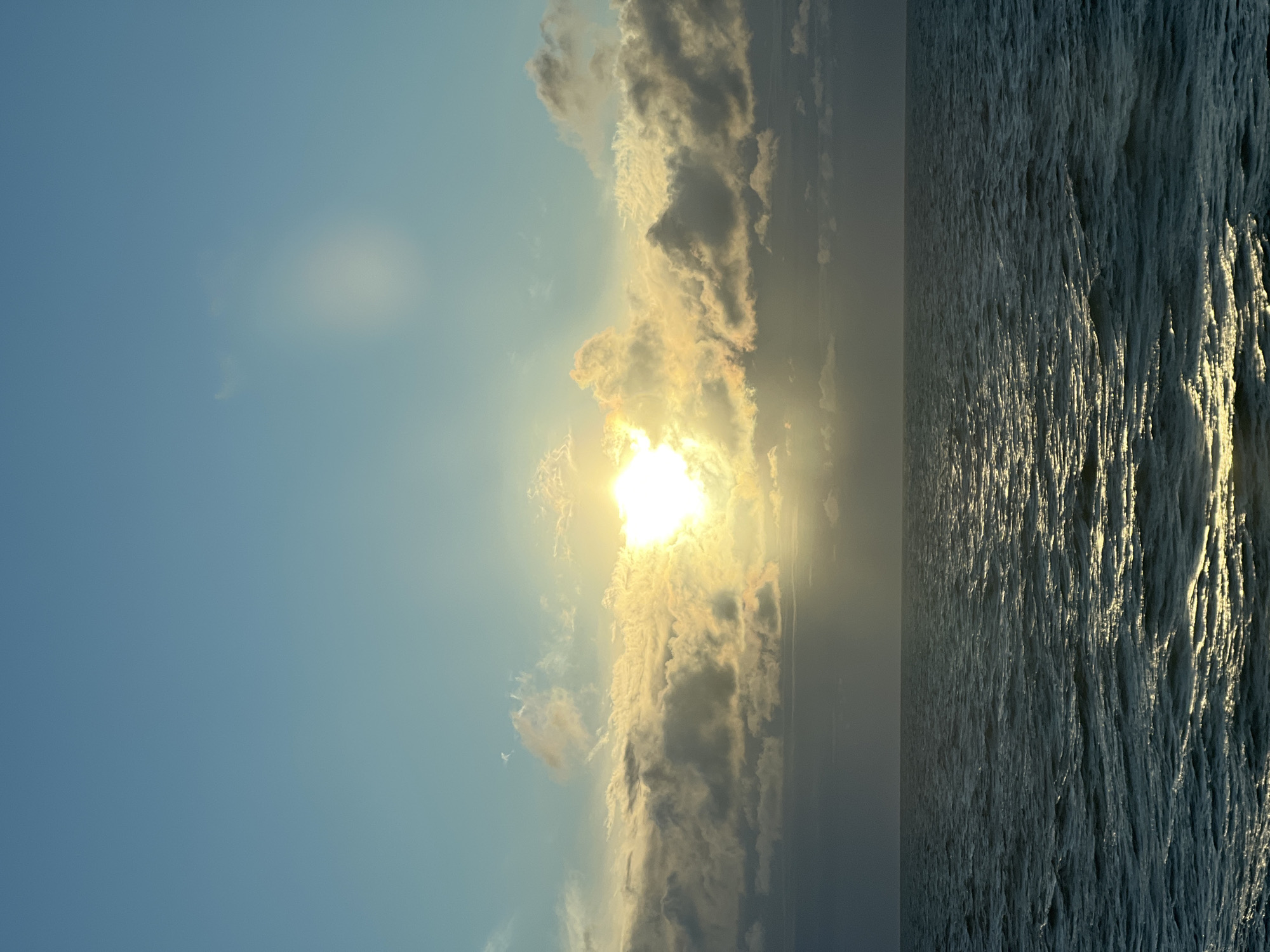 The image depicts a seascape with a clear blue sky above, transitioning to a band of clouds illuminated by the sun. The sun itself is partially obscured by these clouds, creating a bright glow. Below the clouds, the vast expanse of dark, choppy ocean stretches to the horizon. A shimmering reflection of the sun is visible on the water's surface. There are no people or specific structures visible in the frame, suggesting an open water environment. The scene appears to be a natural landscape.