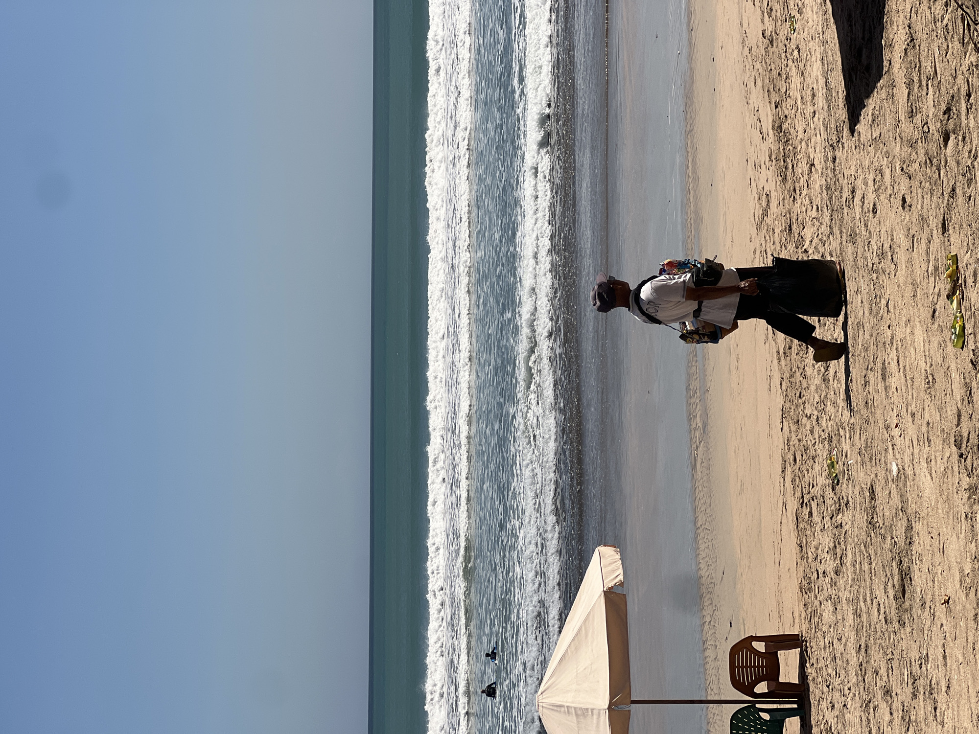 A person walks along a sandy beach in Kuta, Indonesia, carrying a large black bag and a smaller bag slung over their shoulder. The person wears a gray cap, a white t-shirt, and dark pants. In the background, the ocean has moderate waves with whitecaps, and two individuals are visible in the water. To the left of the frame, a beige beach umbrella stands over two empty chairs, one green and one brown. The sand is textured with footprints and debris. The sky is clear and blue.