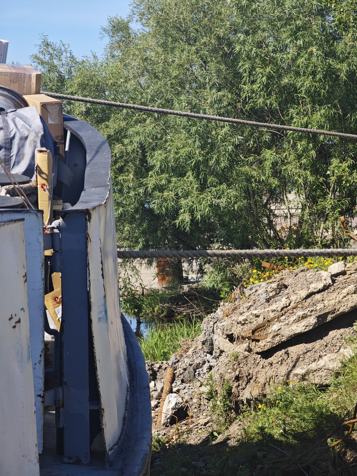The image depicts a close-up view of what appears to be cargo on a vessel or industrial site, with a natural environment in the background. In the foreground, a large, curved, metallic structure, possibly part of a ship's hull or industrial equipment, is visible on the left. It is painted in muted colors, showing signs of wear and rust. Stacked on top of this structure are several cardboard boxes, some appearing to be wrapped in plastic, along with a gray canvas bag. Wires and ropes are also present, connecting to or running alongside the metallic structure.
Behind this industrial element, a dense cluster of green trees with thin leaves dominates the midground and background. A thick, dark rope or cable stretches horizontally across the frame, passing in front of the trees. In the lower right portion of the image, a pile of debris, including large chunks of concrete, rocks, and rusted metal, is visible. Small yellow wildflowers are scattered amongst the green grass growing around the debris. A narrow strip of water, reflecting the blue sky, can be seen between the industrial structure and the debris pile. The sky above is a clear, bright blue.
The scene is outdoors, under daylight, suggesting it is a sunny day. No people are visible in the image. The overall impression is one of a functional, perhaps slightly disused, industrial area adjacent to a natural landscape. The presence of cargo and industrial elements suggests a possible port or logistics hub, while the trees, grass, and water point to a location near a natural body of water or a park.
Without further context or identifiable landmarks, it is impossible to definitively confirm the location as Krasnoyarsk, Russia. The image provides a snapshot of a specific scene that could exist in various industrial or port areas globally.