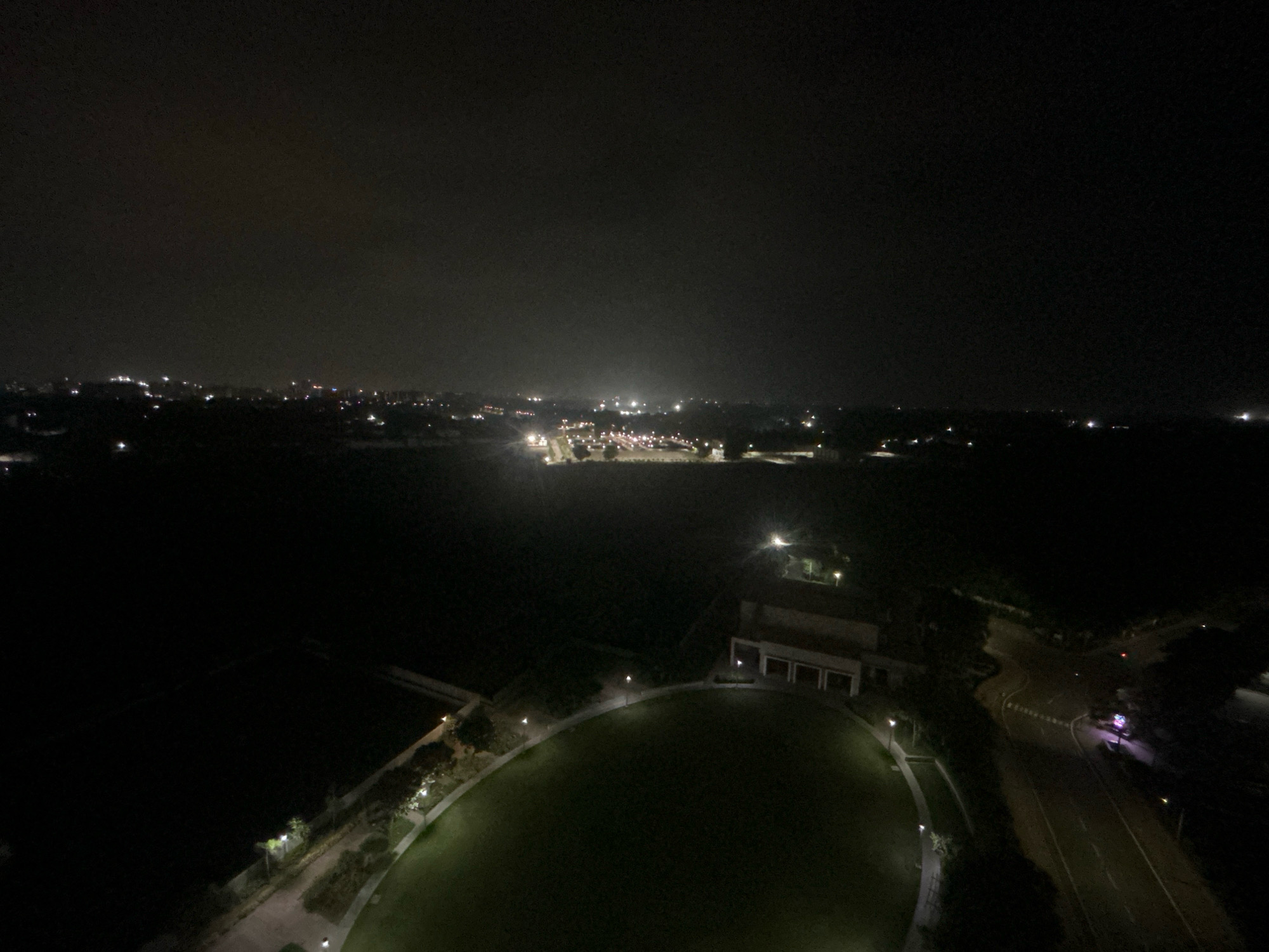 This is a nighttime aerial photograph of a developed area in Shantigram, India. The scene is dominated by darkness, with artificial lights illuminating scattered structures and roadways. In the foreground, a large, dark, circular green space, possibly a park or a stadium, is visible. Surrounding this area are paved walkways and scattered trees, all lit by intermittent lampposts. To the right of the green space, a building complex with a light-colored facade and multiple arched entrances is illuminated. Adjacent to the building and walkways is a road that curves through the landscape, with yellow and white lines marking its lanes. Further in the distance, a city skyline is visible, characterized by numerous clusters of bright lights indicating a populated urban area. The sky is dark and hazy, suggesting low visibility or a cloudy night. No individuals are discernible in the image.