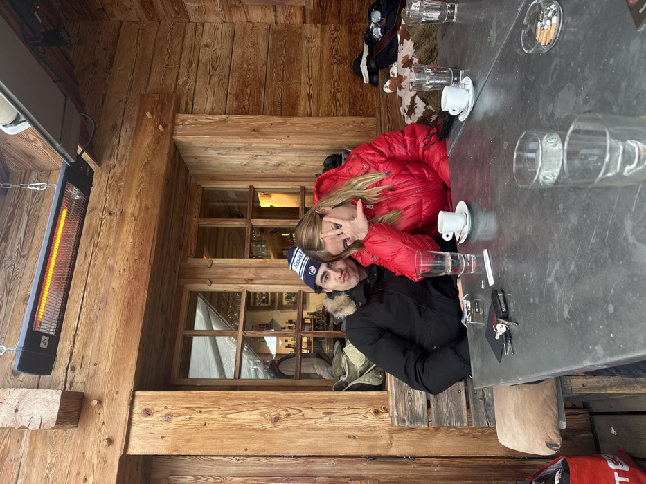 This image depicts two individuals seated at a grey table on an exterior patio of a wooden chalet-style building in La Clusaz, France. An infrared heater is suspended overhead, emitting an orange glow. The male on the left, wearing a black puffer jacket with a fur-trimmed hood and a blue, white, and red "Risal" beanie, is looking towards the camera with pursed lips. To his right, a female with blonde hair, dressed in a red puffer jacket, also looks towards the camera with her right hand raised, fingers splayed, partially obscuring her face.
On the table, from left to right, are a pack of Winston cigarettes, a set of keys on a dark object, a tall glass filled with water, and a white espresso cup with its saucer. Further right on the table, another white espresso cup with its saucer, two additional tall glasses (one partially filled with water), and an ashtray containing multiple cigarette butts are present. In the background, a large multi-paned window reveals an interior space with shelves of bottles and glasses. To the far right, on a wooden bench, ski goggles and a brown and white patterned blanket are visible. The entire structure is constructed from light-colored wooden planks and beams.