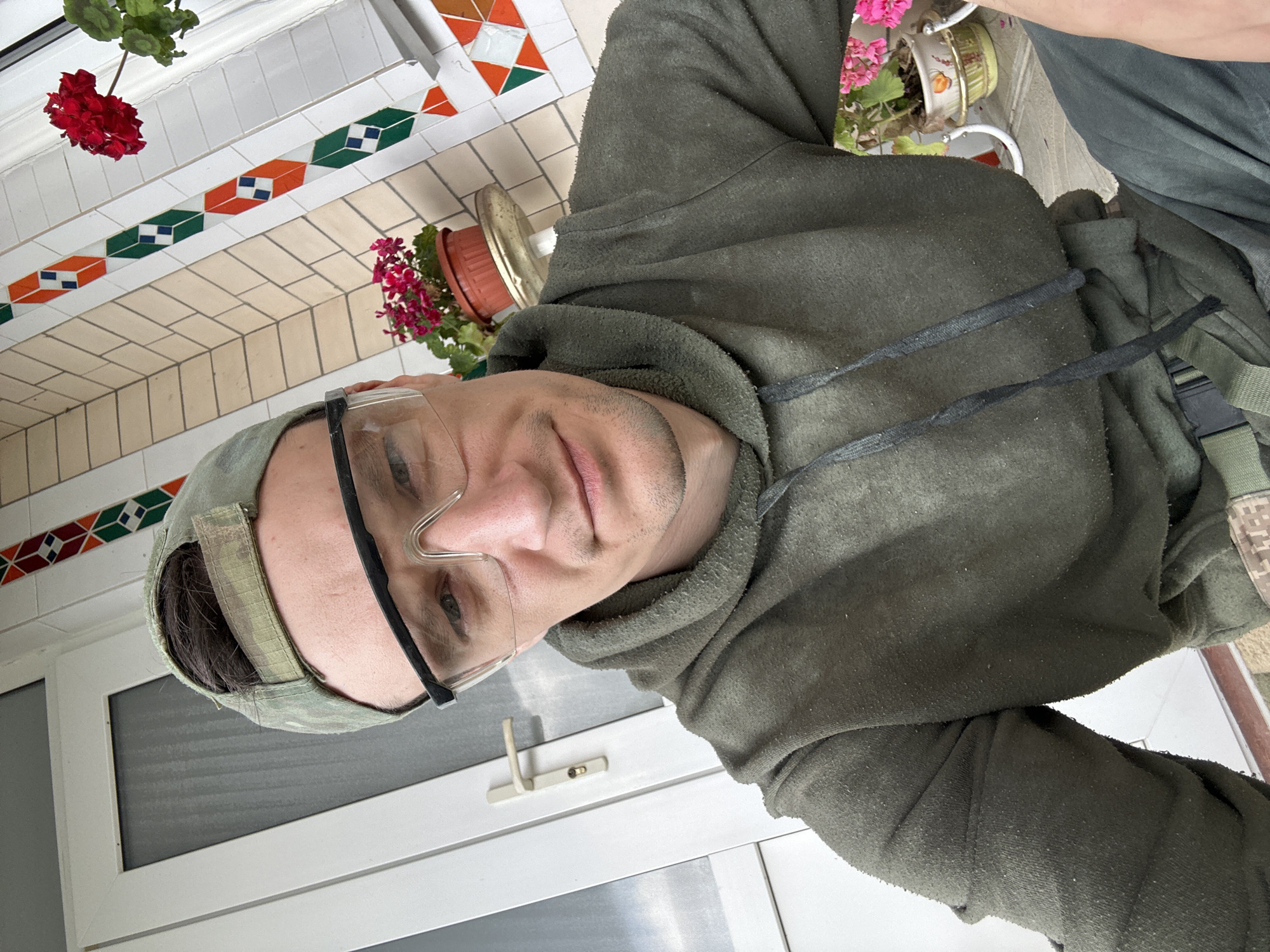 The image shows a close-up, front-facing selfie of a person wearing a green camouflage baseball cap, clear protective eyewear, and a dark green hoodie. The person has short, dark hair and a light complexion, with a slight smile. The background suggests an outdoor setting, possibly a porch or patio area. To the left, a white door with a silver handle is visible. To the right, a tiled wall with a decorative pattern of colorful geometric shapes and potted plants with pink and red flowers can be seen. One of the plants has a red flower and is placed on a small, ornate stand. The overall impression is casual, with the subject possibly engaged in some outdoor activity given the protective eyewear. The specific location in Makiv, Ukraine is not discernible from the visual information provided in the image.