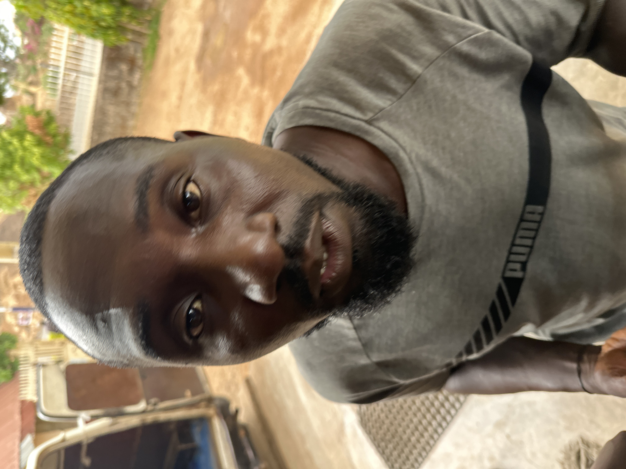 A close-up, slightly angled photograph captures a dark-skinned man with a trimmed beard, wearing a grey Puma t-shirt with a black stripe, and positioned outdoors. The background reveals a glimpse of urban scenery, with vegetation, buildings, and what appears to be a vehicle, suggesting an outdoor environment within a city, possibly Jos, Nigeria.