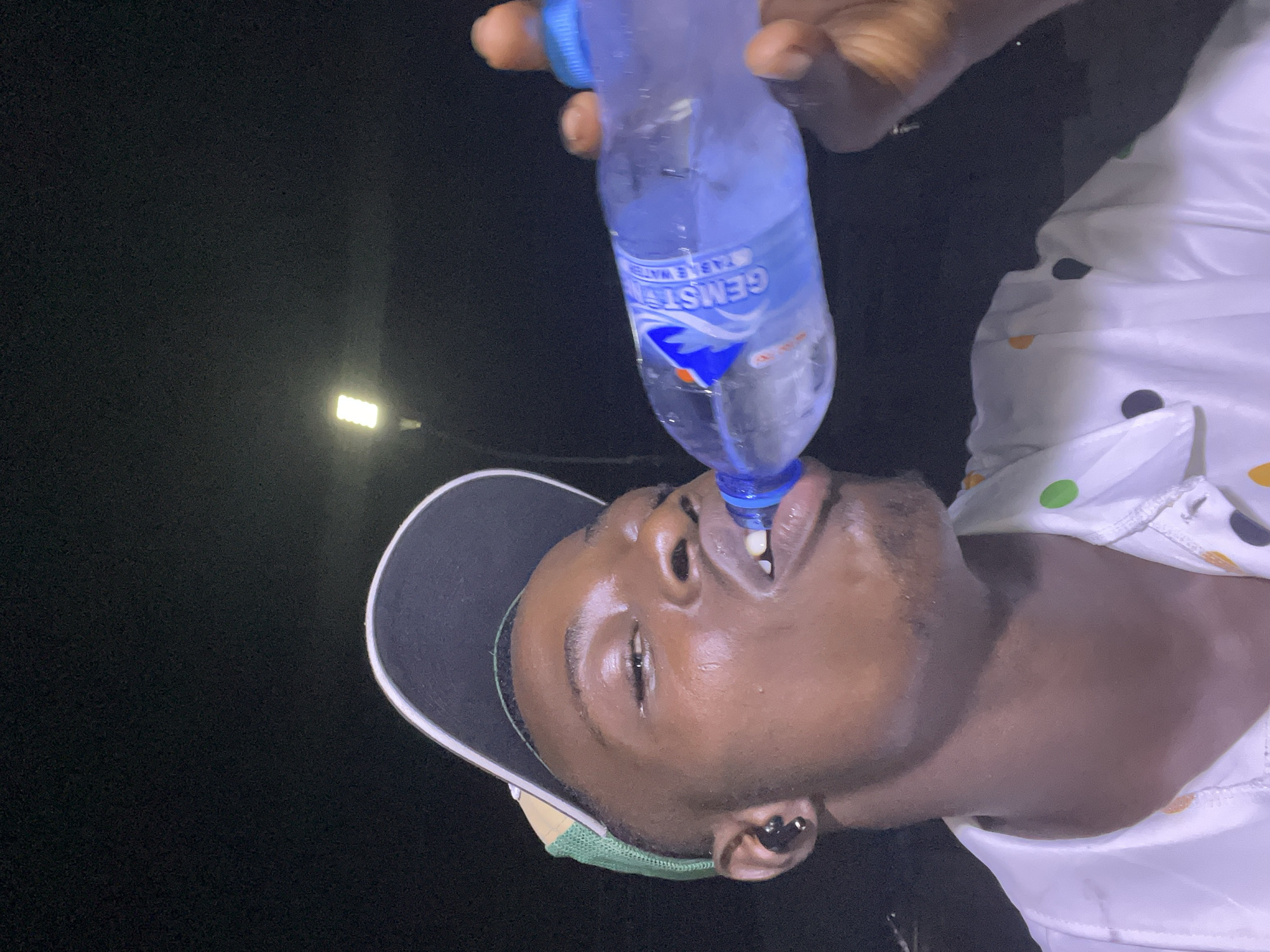 A man is drinking from a bottle of Gemstar table water at night. He is wearing a white shirt with colorful polka dots, a baseball cap, and an earbud. He is standing outdoors, with a street light visible in the background. The image appears to have been taken in Nkpor, Nigeria.