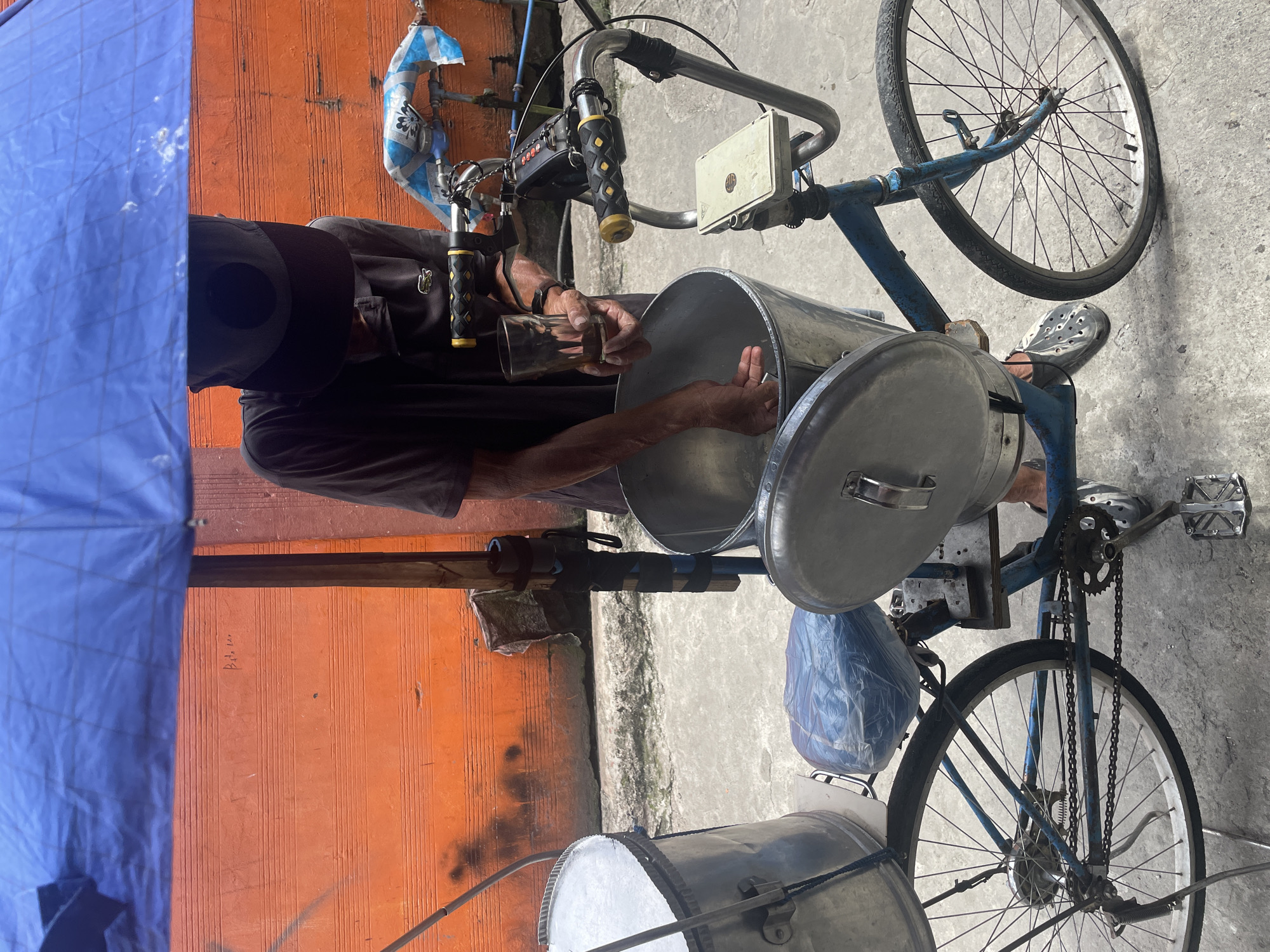A person, partially obscured by a blue tarp and a baseball cap, stands next to a modified bicycle in what appears to be a street in Davao City, Philippines. The bicycle has been outfitted with two large metal containers, one in front and one behind the seat, suggesting it's used for mobile vending. The person is holding a glass and appears to be serving from the front container, which is open. The background features a textured orange wall. The ground is paved.