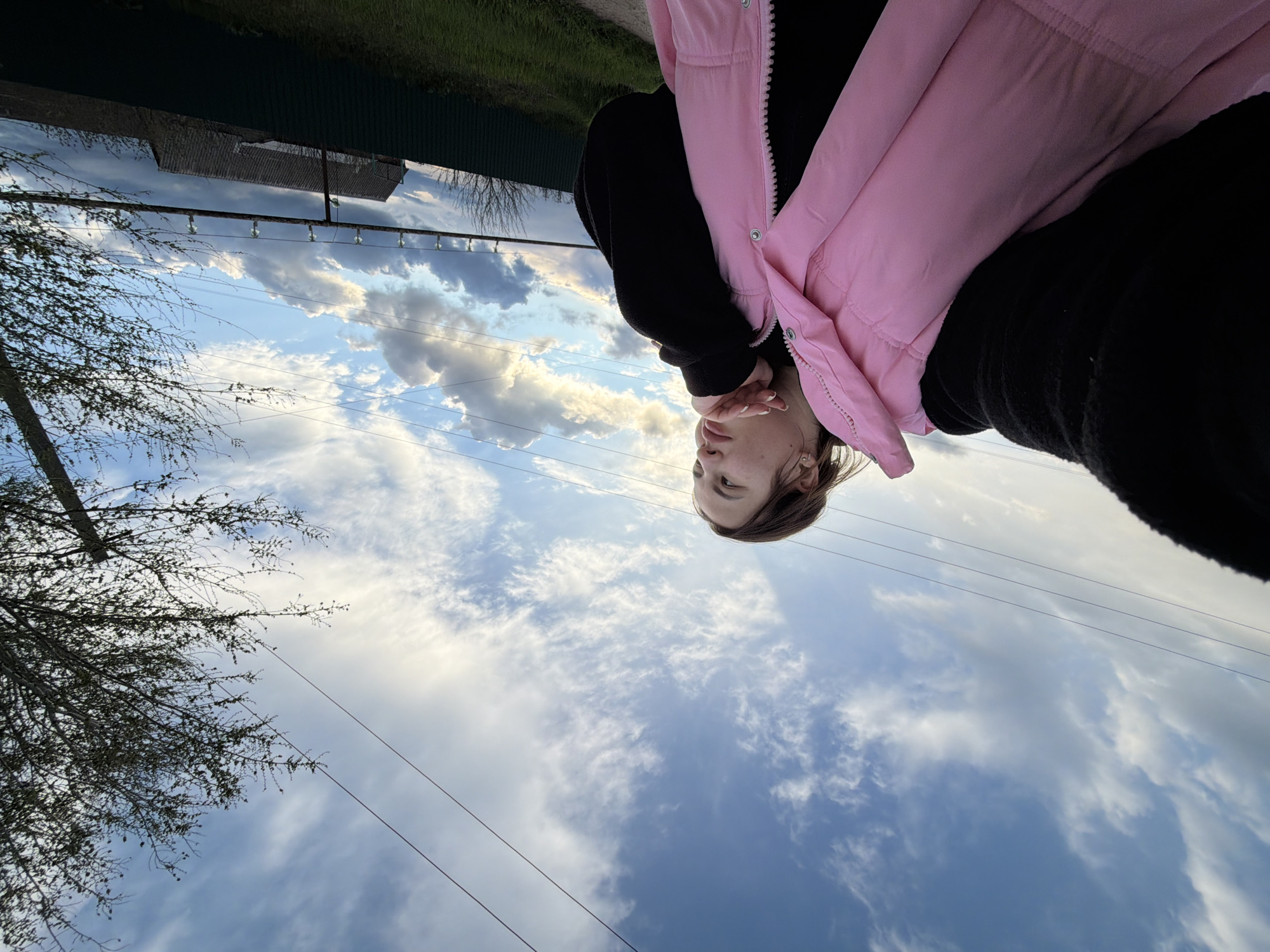 The image is an upside-down view of a person wearing a pink vest and black clothing, with the sky and clouds as the primary backdrop. The location appears to be outdoors, possibly in a residential area, as indicated by a building roof and power lines visible in the background. There's also foliage from a tree visible on the left side. The person's face is partially visible, with their hand near their face. The exact location within the city of Unknown, Unknown cannot be determined from the image.