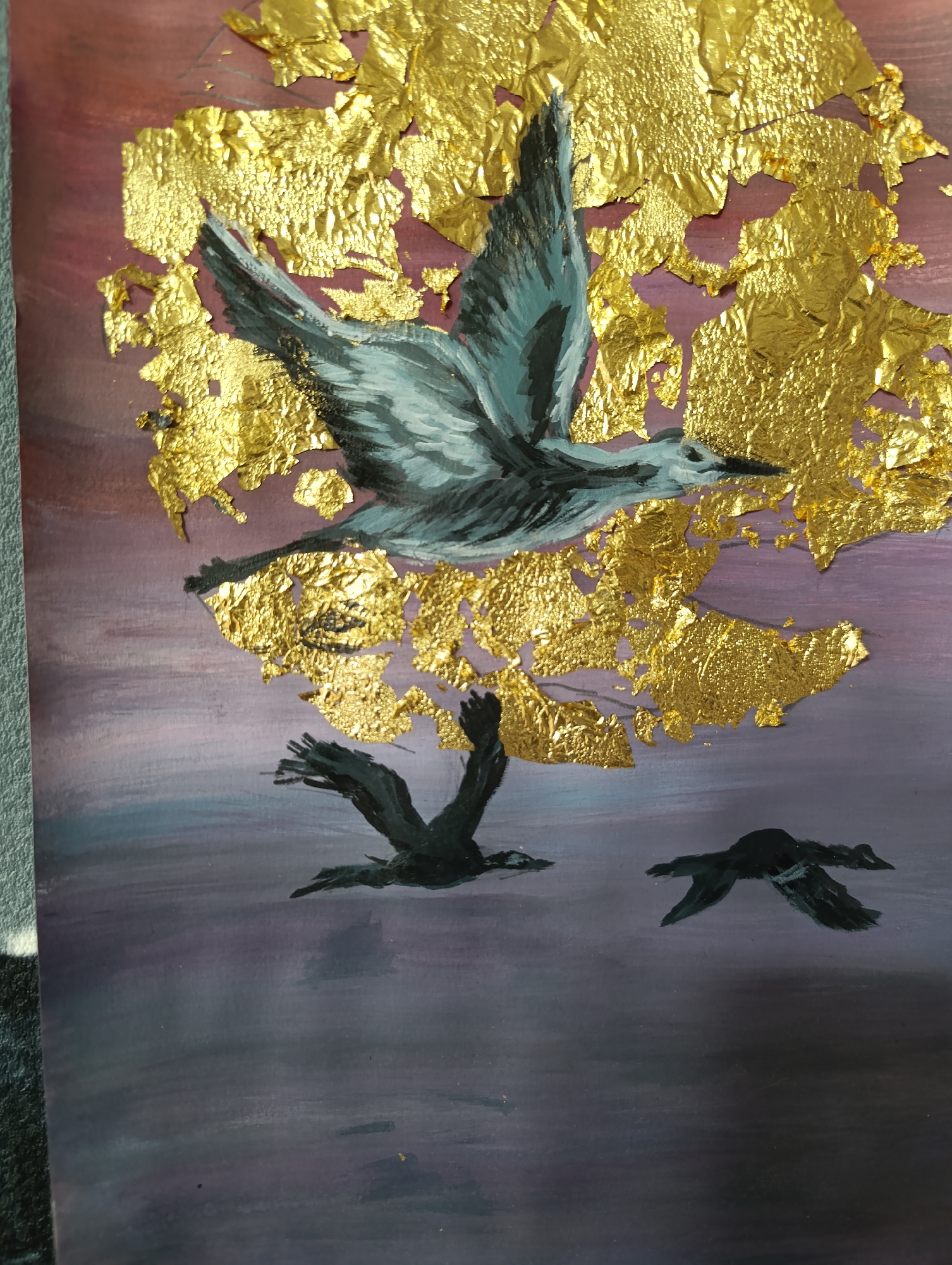 This is a mixed-media artwork depicting birds in flight against a gradient sky, and as an artwork, it is not situated in a real-world city of Unknown, Unknown.
The scene features three birds in various stages of flight. The background transitions from a reddish-pink hue in the upper left, blending into purples and lavenders through the mid-section, and darkening to deep indigo and muted grey-blue tones in the lower two-thirds, suggesting a twilight or pre-dawn sky or reflective water surface.
Prominently featured in the upper-middle section is a large bird with its wings fully outstretched upwards, appearing to ascend. This bird is painted in shades of grey with lighter white highlights on its wings and belly, and darker accents on its head and tail.
Extensive irregular patches of crumpled, reflective gold leaf are applied across the upper two-thirds of the artwork, concentrated around and above the main bird, creating a textured, shimmering effect that contrasts sharply with the painted background. Smaller, scattered flakes of gold leaf trail downwards from the main concentration.
In the lower half of the painting, two smaller, silhouetted birds are depicted in flight, rendered in dark black or very dark grey tones. One is positioned towards the lower left, and the other further to the right. Both have their wings extended horizontally, appearing to fly closer to the darker background. All three birds are generally oriented as if moving from left to right across the canvas.