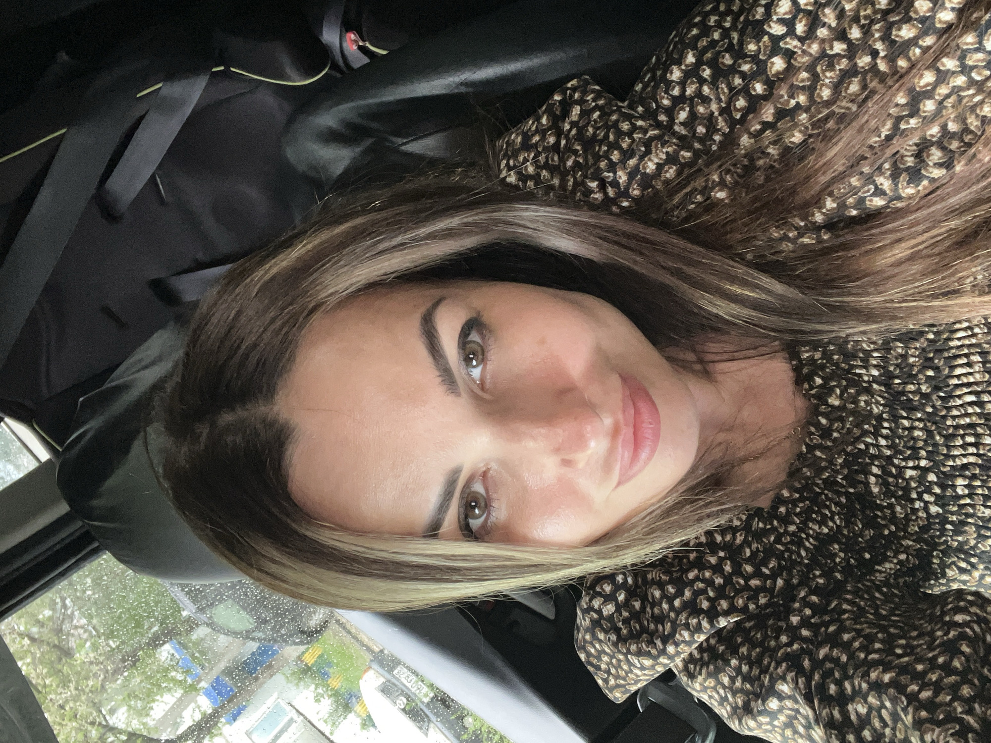 A woman with brown hair is taking a selfie in a car. She is wearing a patterned shirt. The car interior is visible, as is the outdoors through the window.