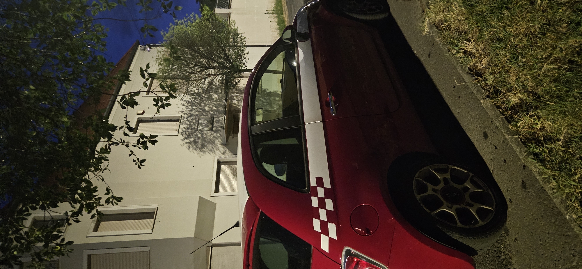 A red Fiat 500 hatchback is parked on a curb at night. The car has a white stripe along its side and a checkerboard decal on its rear quarter panel. The car is parked in front of a light-colored building with windows covered by blinds. Trees are visible in front of the building, casting shadows on the facade. The sky is dark blue, indicating nighttime. The scene appears to be in Hévíz, Hungary, based on the surrounding environment.