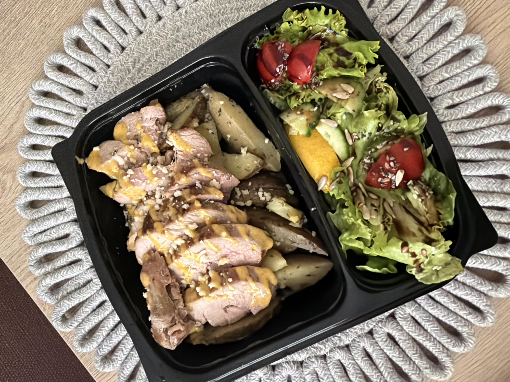 A top-down view shows a black compartmentalized take-out container filled with food, placed on a textured grey placemat. The container holds sliced meat with a yellow sauce and crumbled cheese, alongside potato wedges in the upper compartment. The lower compartment contains a salad with lettuce, tomatoes, cucumber, a slice of orange, and various seeds, drizzled with dressing. The background is a wooden surface. The image does not contain any information that could be used to determine a location in Pazardzhik, Bulgaria.