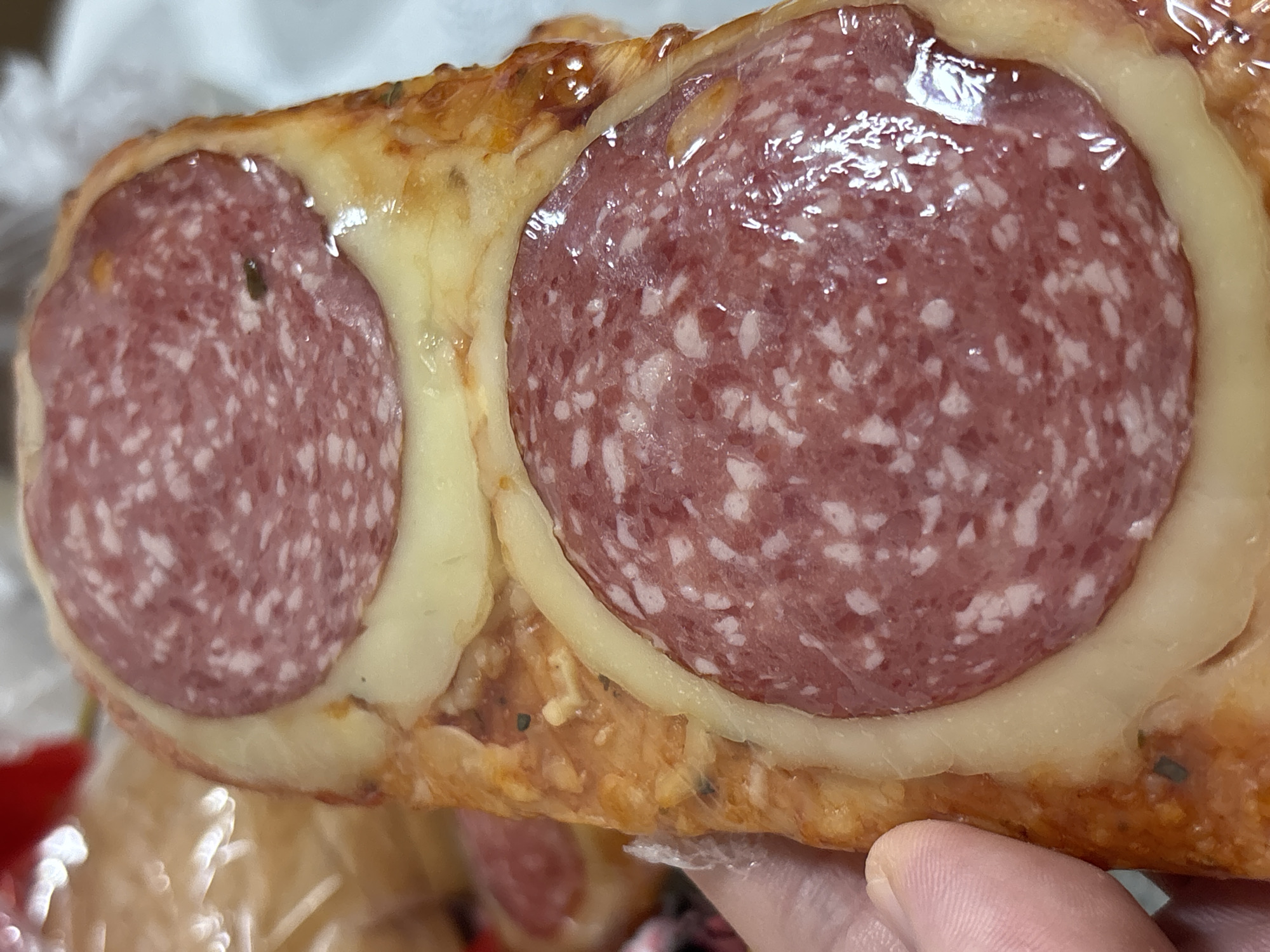 The image shows a close-up of a food item, possibly a piece of pizza or a baked pastry, topped with slices of salami and melted cheese. Two prominent slices of salami are visible, embedded in a golden-brown crust with red sauce and melted cheese. The salami slices are circular with a characteristic marbled appearance of cured meat. A person's thumb is visible on the left side of the image, indicating that the food item is being held. In the background, out of focus, there appears to be some packaging, possibly plastic wrap, and other food items. The lighting suggests an indoor setting. There is no information in the image that would indicate a location in Ostrov, Russia.