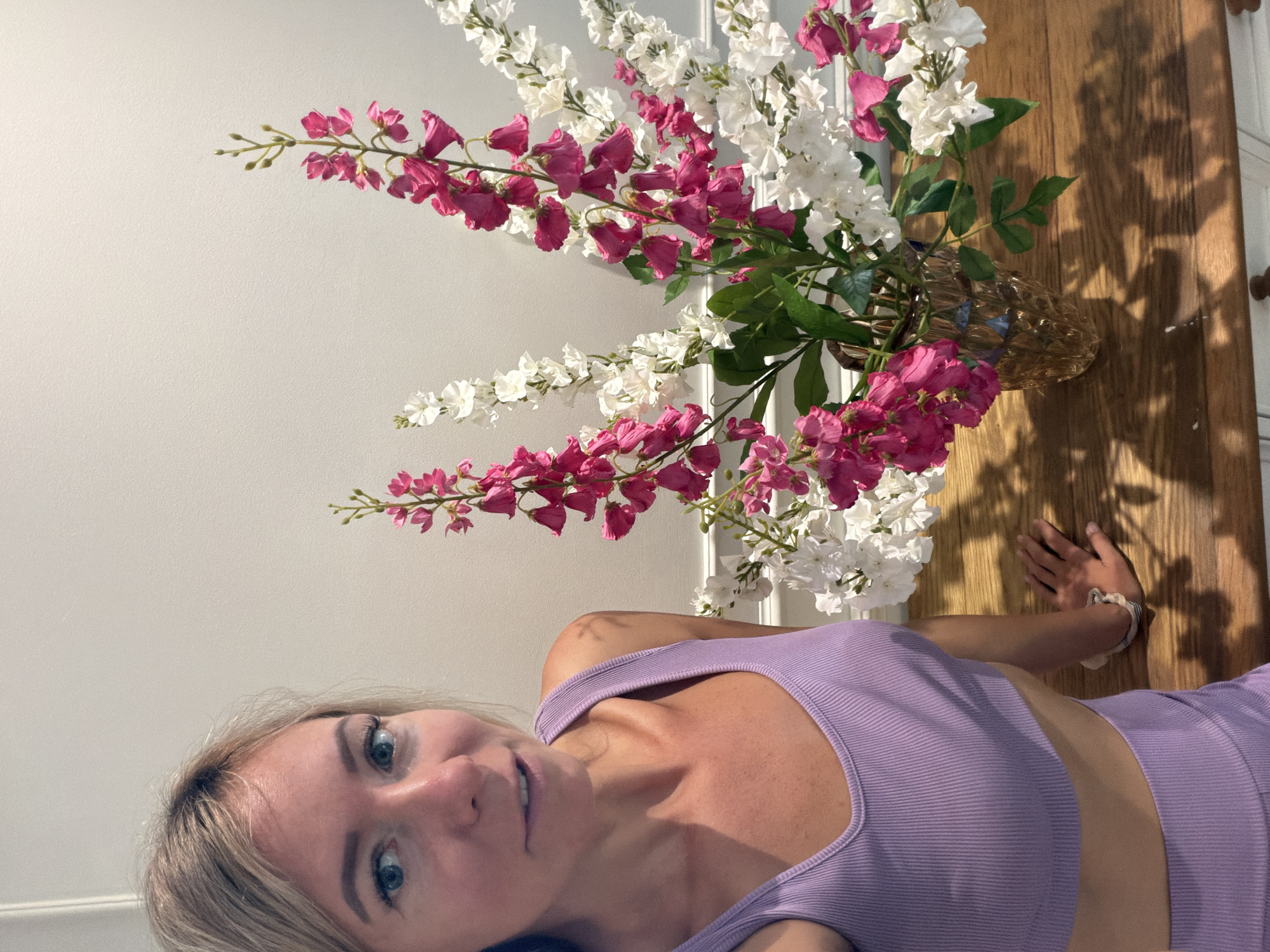 A woman with blonde hair and blue eyes is sitting at a wooden table, with a vase of pink and white flowers to her right. She is wearing a purple sports bra and matching shorts. The wall behind her is white. The scene appears to be indoors, possibly in a home. The location is Xuân Trường - Đà Lạt, Vietnam.