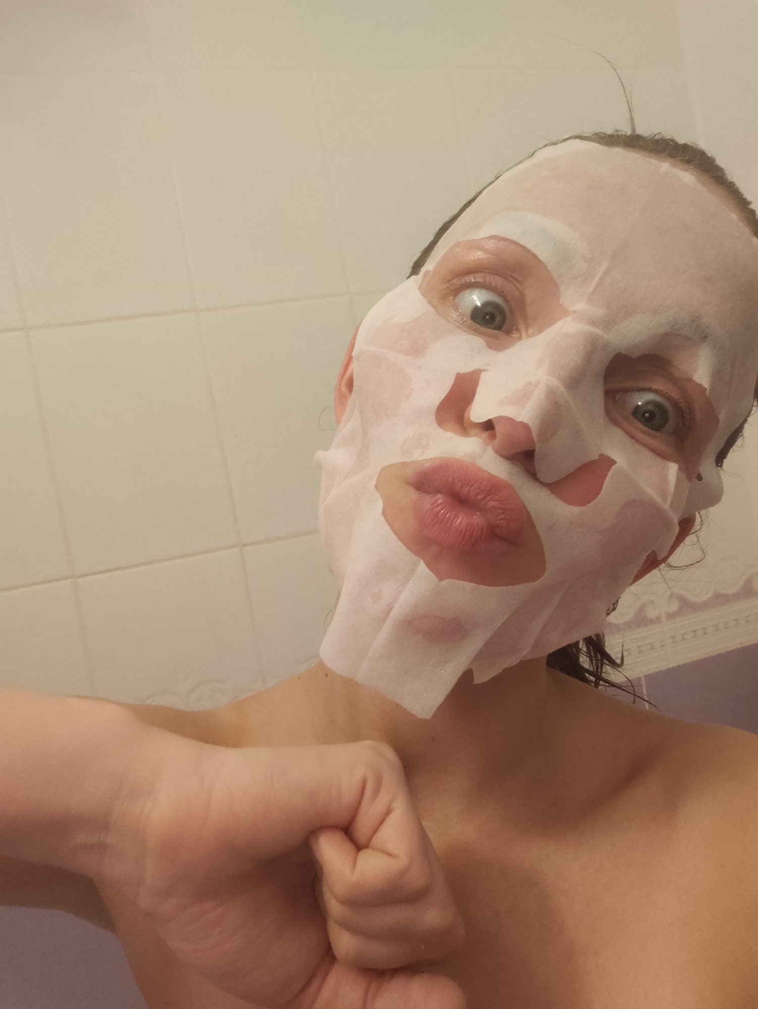 A woman is taking a selfie in a bathroom. She is wearing a white sheet mask on her face and pursing her lips. Her hair appears to be wet. She is making a fist with her left hand, which is positioned in the foreground. The background consists of white bathroom tiles. The location is potentially a residence in Chaikovsky, Russia.