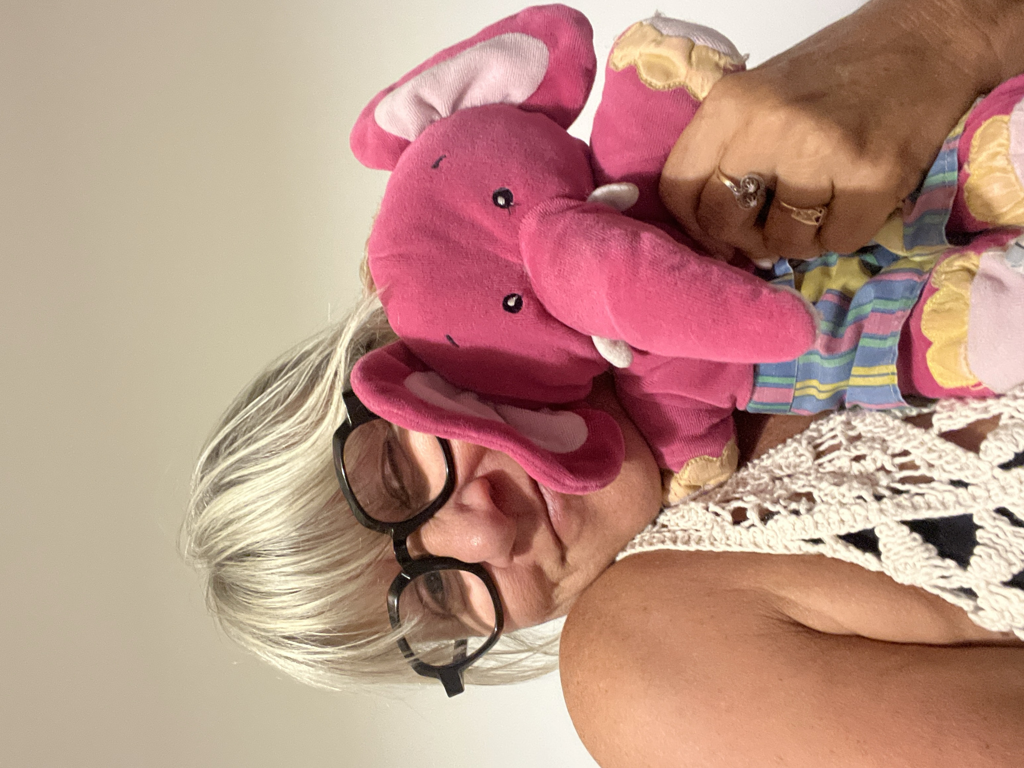 The image shows a close-up, waist-up shot of a woman holding a pink stuffed elephant. The woman has short, blonde hair, wears large, black-rimmed glasses, and is looking towards the camera with one eye closed in a wink. She is wearing a white crocheted top over a dark garment. The stuffed elephant is pink with light pink accents on its ears and is wearing striped pants. The woman's hand, which is holding the elephant, wears two rings. The background is a plain, light-colored wall. There is no indication of the city of Chambœuf, France in the image.