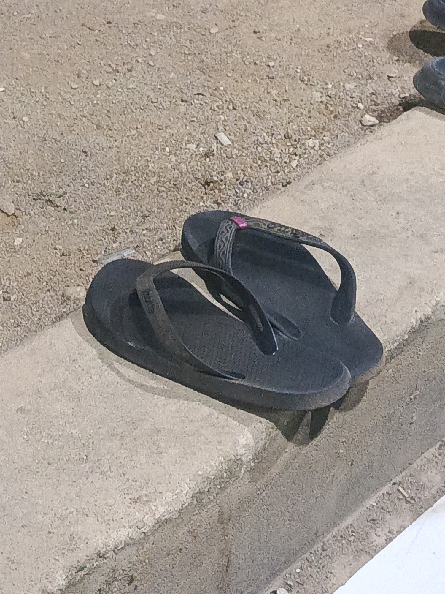 Two black flip-flops are resting on a concrete ledge. The flip-flops are made of rubber and have a textured surface. The left flip-flop is slightly in front of the right one. The ledge is made of concrete and has a rough texture. Behind the ledge is a ground surface covered with small stones and dirt. The scene appears to be outdoors, possibly in a park or a street.