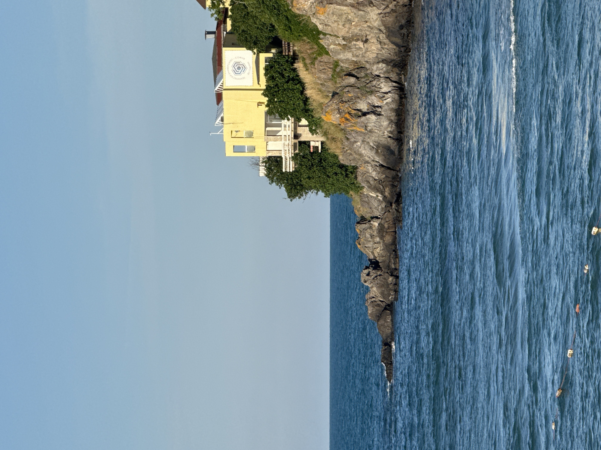 This is a photograph taken in Sozopol, Bulgaria, featuring a yellow building perched on a rocky cliff overlooking the sea. The building appears to be a university or research facility, as evidenced by a logo with Cyrillic text that reads "ТЕХНИЧЕСКИ УНИВЕРСИТЕТ - СОФИЯ" (Technical University - Sofia). The building has multiple windows and balconies, and is surrounded by lush green trees. The sea is a deep blue, with gentle waves breaking against the shore. The sky is clear and blue, suggesting a bright, sunny day. In the water, a series of buoys or markers are visible, connected by a rope. The scene evokes a sense of tranquility and intellectual pursuit, set against the natural beauty of the Bulgarian coast. The photograph is oriented vertically, with the cliff and building occupying the right half of the frame, and the sky and sea filling the left. The lighting indicates it was taken during daylight hours, likely in the morning or afternoon.