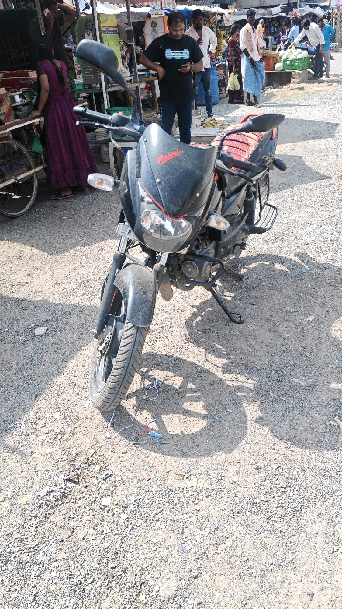 A motorcycle is parked on a gravel road in Pamur, India. The motorcycle is black and has a red seat. A man in a black t-shirt is standing next to the motorcycle, looking at his phone. A woman in a purple dress is standing behind the motorcycle, looking at a stall. There are other people and stalls in the background. The scene appears to be a market.