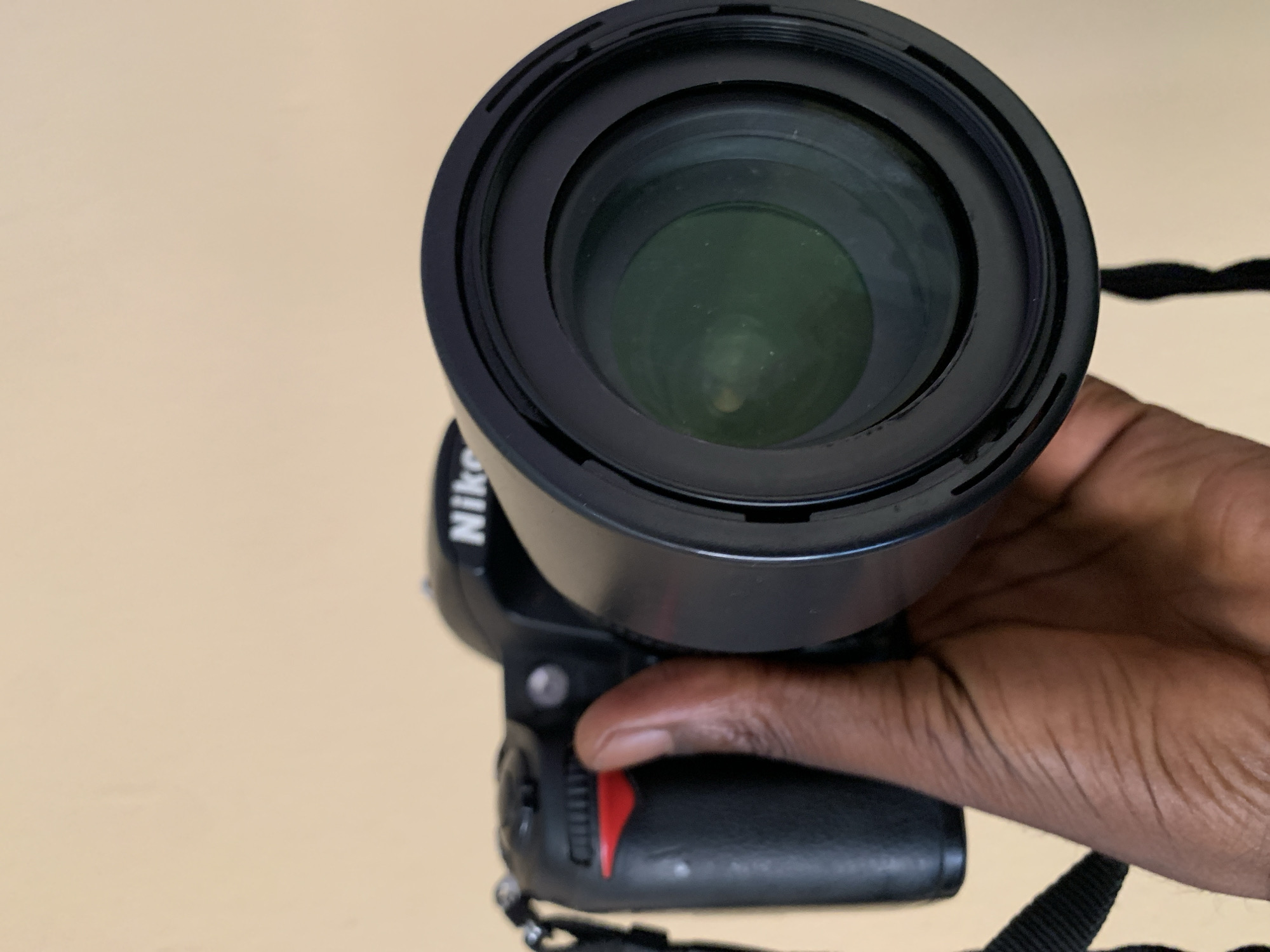 A close-up shot shows a hand holding a black Nikon camera. The camera is pointed downwards, with the lens facing the viewer. The hand's dark skin is visible, with its fingers wrapped around the camera's grip and thumb resting on a red dial. The camera itself has a prominent black lens barrel with concentric rings. The Nikon logo is partially visible on the camera body. The background is a plain, light-colored surface, possibly a table or wall, providing no specific location cues. The lighting suggests an indoor setting, likely during the day, as there are no harsh shadows. The overall impression is a neutral, unposed shot focused on the camera and the act of holding it. There are no discernible weather or emotional cues. The visible text is limited to the "Nikon" branding on the camera.