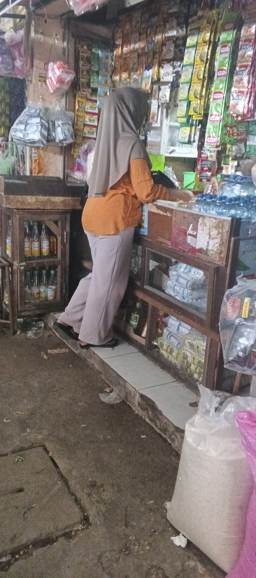 A woman wearing a grey hijab and an orange shirt is standing in a small, cluttered shop. She is facing a counter and appears to be interacting with the items on it, which include bottled drinks and packaged goods. The shop is filled with shelves of various products, some hanging from the ceiling and others displayed in wooden cabinets. The floor is made of rough concrete, and there are some plastic bags on the ground. The scene suggests a typical day at a local convenience store in Sekaran, Indonesia, where such small businesses are common for daily necessities.