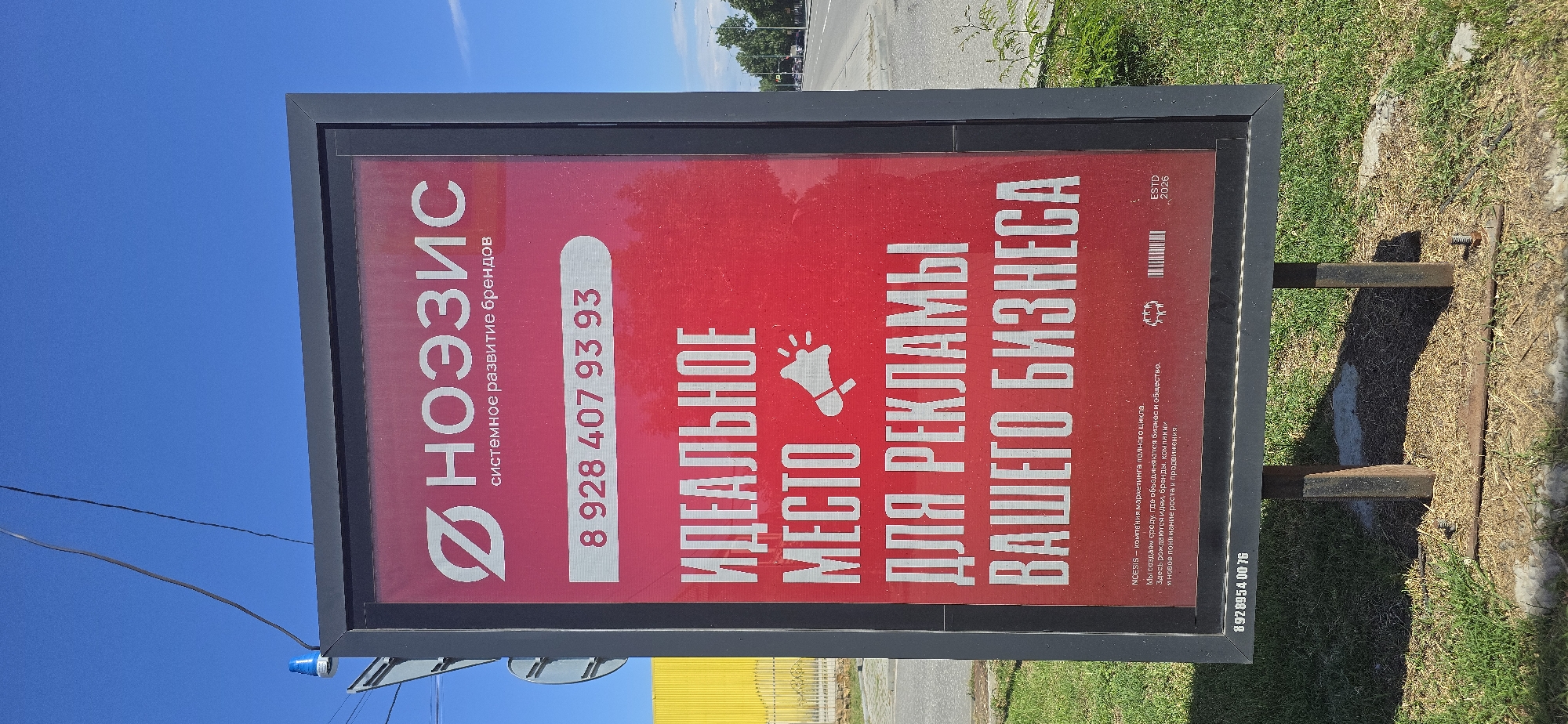 A billboard is displayed outdoors with a bright red background. The billboard features white text and a logo. At the top, the logo consists of a stylized symbol and the Russian word "НОЭЗИС" (NOEZIS). Below this, in smaller text, it says "системное развитие брендов" (systematic brand development). A phone number, "8 928 407 93 93", is prominently displayed in the center. Below the phone number, large white text reads "ИДЕАЛЬНОЕ МЕСТО ДЛЯ РЕКЛАМЫ ВАШЕГО БИЗНЕСА" (IDEAL PLACE FOR YOUR BUSINESS ADVERTISING). There is also a small graphic of a megaphone next to the word "МЕСТО". At the bottom of the billboard, smaller text provides a description of the company, followed by icons and "ESTD 2026". A smaller text at the very bottom left appears to be a phone number "8928954 00 76". The billboard is supported by two metal posts and is situated in a grassy area. In the background, a yellow building is visible, as well as trees, a street, and cars. The sky is clear and blue. The image is taken from a slightly low angle, looking up at the billboard. The location is identified as Grozny, Russia.