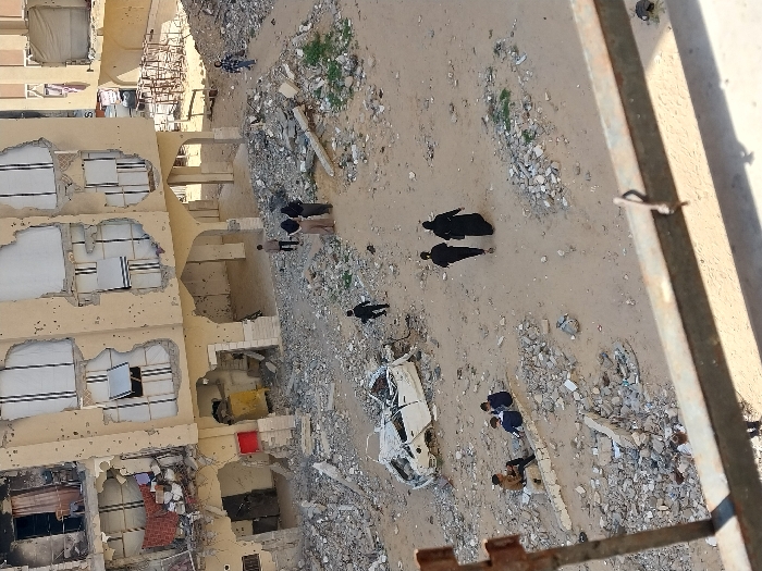 The image depicts a devastated urban landscape in Khan Yunis, Palestinian Territory, characterized by extensive structural damage and debris. In the foreground, a rusty metal railing from a vantage point partially obscures the lower edge of the frame. The ground is a mix of sand and compacted dirt, heavily covered with rubble, shattered concrete fragments, and other debris.
The dominant feature is a multi-story building, likely residential or mixed-use, in the center-left to background. Its light-colored exterior walls are pockmarked with numerous small holes, indicative of bullet impacts or shrapnel. Large sections of the facade are blown out, revealing damaged interior rooms. Windows are either destroyed, missing, or haphazardly covered with white panels. The ground floor features several arched openings, now largely filled with debris. To the right of this building, further structures appear similarly damaged, and a skeletal metal frame or structure is visible.
Numerous individuals are present in the scene. In the lower-left, three males, appearing to be youths, sit on a low, damaged concrete structure amidst the rubble. One wears a brown top, and the other two wear dark blue. To their right, a white, heavily damaged and crumpled vehicle, possibly a sedan, lies overturned or severely crushed amidst the debris. Further right, a single child in dark clothing walks across the sandy ground. Three adults walk together in the mid-ground, heading towards the lower-right; two are in dark attire, one appearing to wear a full abaya, and the third is a male in a light top and dark trousers. Closer to the main damaged building, three other individuals stand near its ground-floor arches. In the far background-right, an additional individual walks near another damaged structure. The sky above is clear and bright.