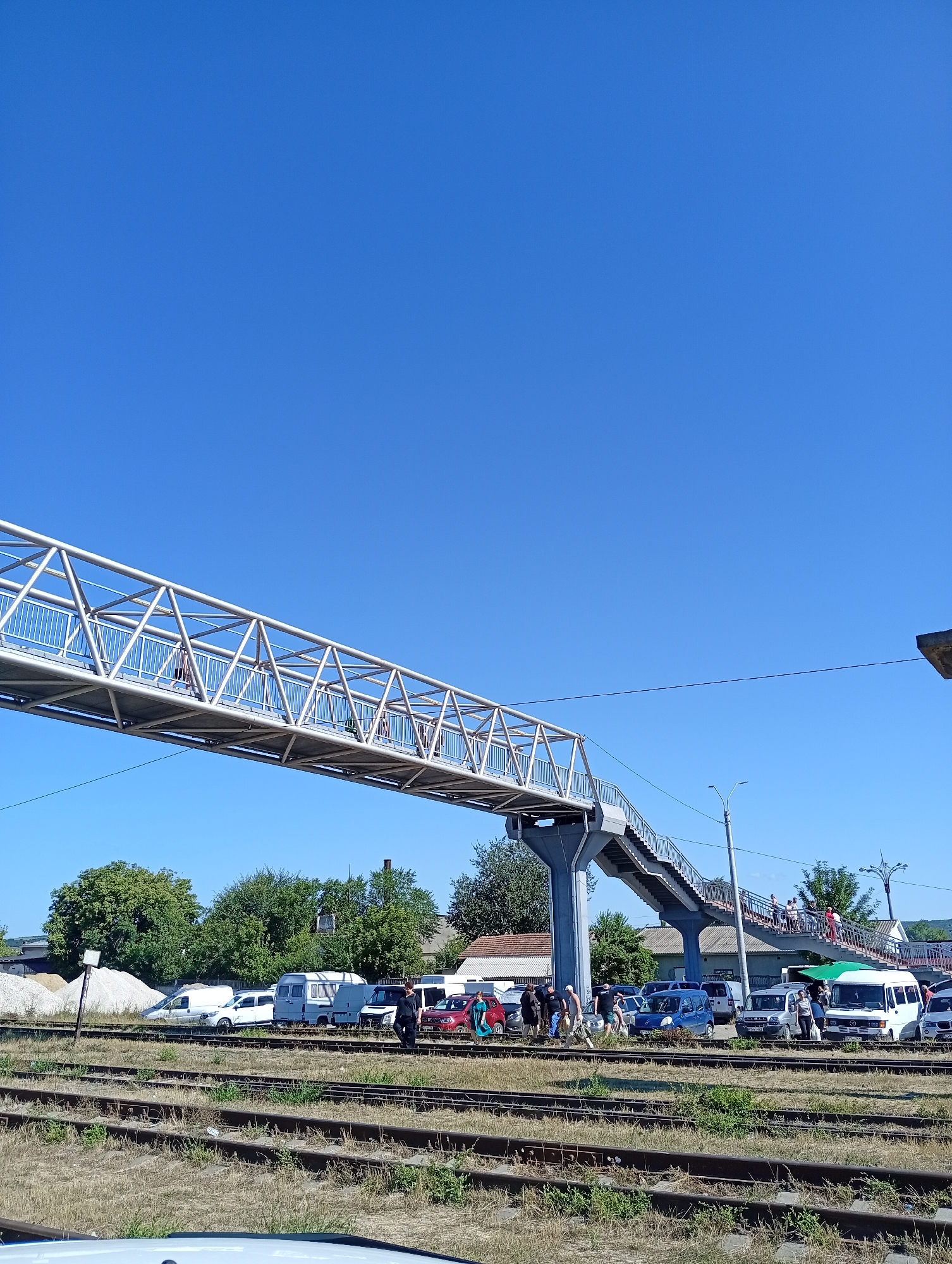 In Strășeni, Moldova, on a clear, sunny day, a pedestrian overpass spans multiple railway tracks. The overpass is a silver, metal structure with a walkway and stairs. Several people are visible walking on the overpass and descending the stairs. Below the overpass, on the other side of the railway tracks, a variety of vehicles are parked, including cars, vans, and a white minibus. A pile of white gravel is situated to the left of the parked vehicles, and various trees and buildings are visible in the background. The railway tracks are bordered by dry grass.