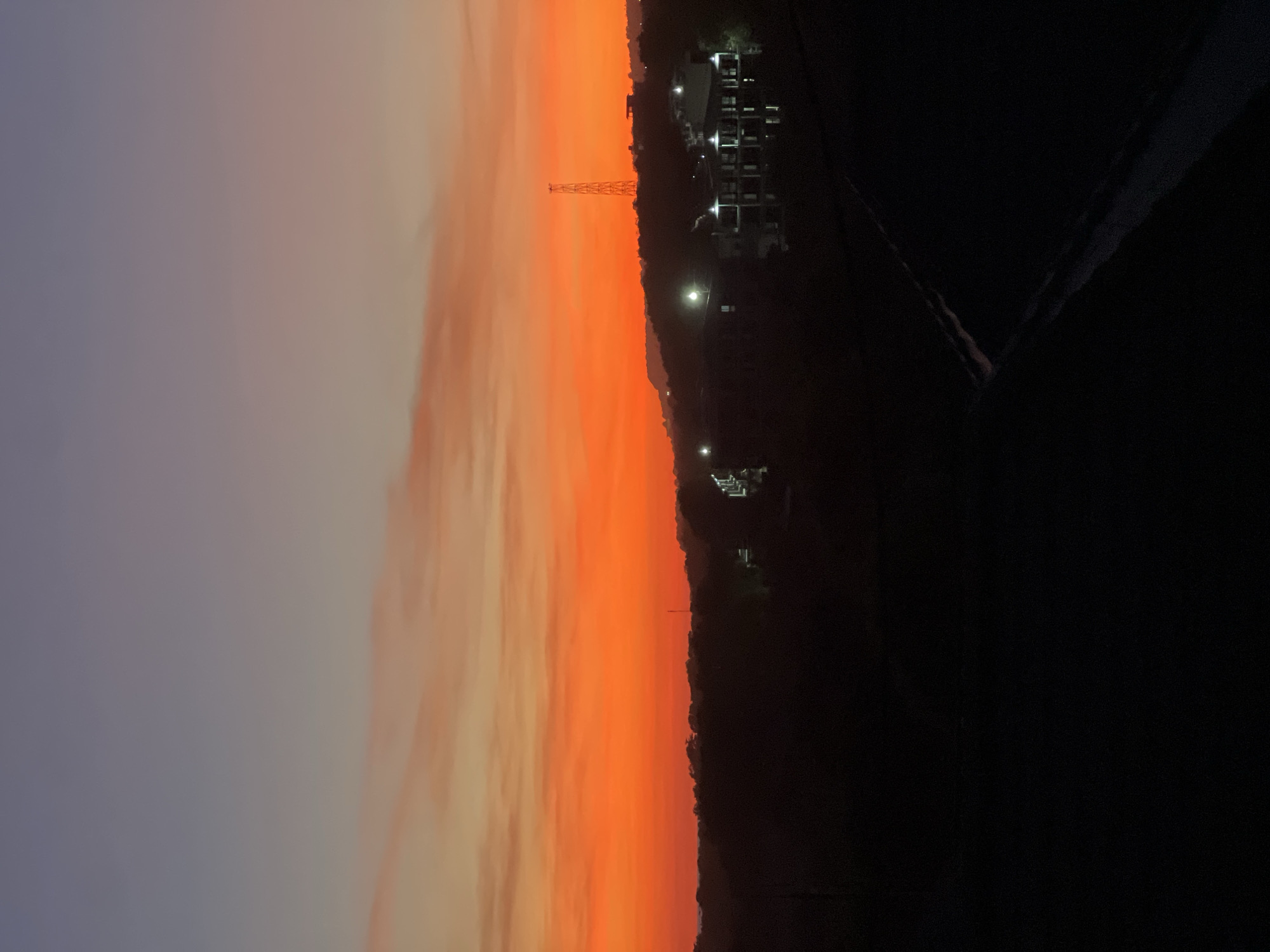 The image depicts a sunset over a city. The sky is predominantly orange, with hints of purple and gray. On the right side of the frame, buildings with illuminated windows are visible, along with a communication tower. The foreground is mostly dark, with indistinct shapes. The location is Unknown, Unknown.