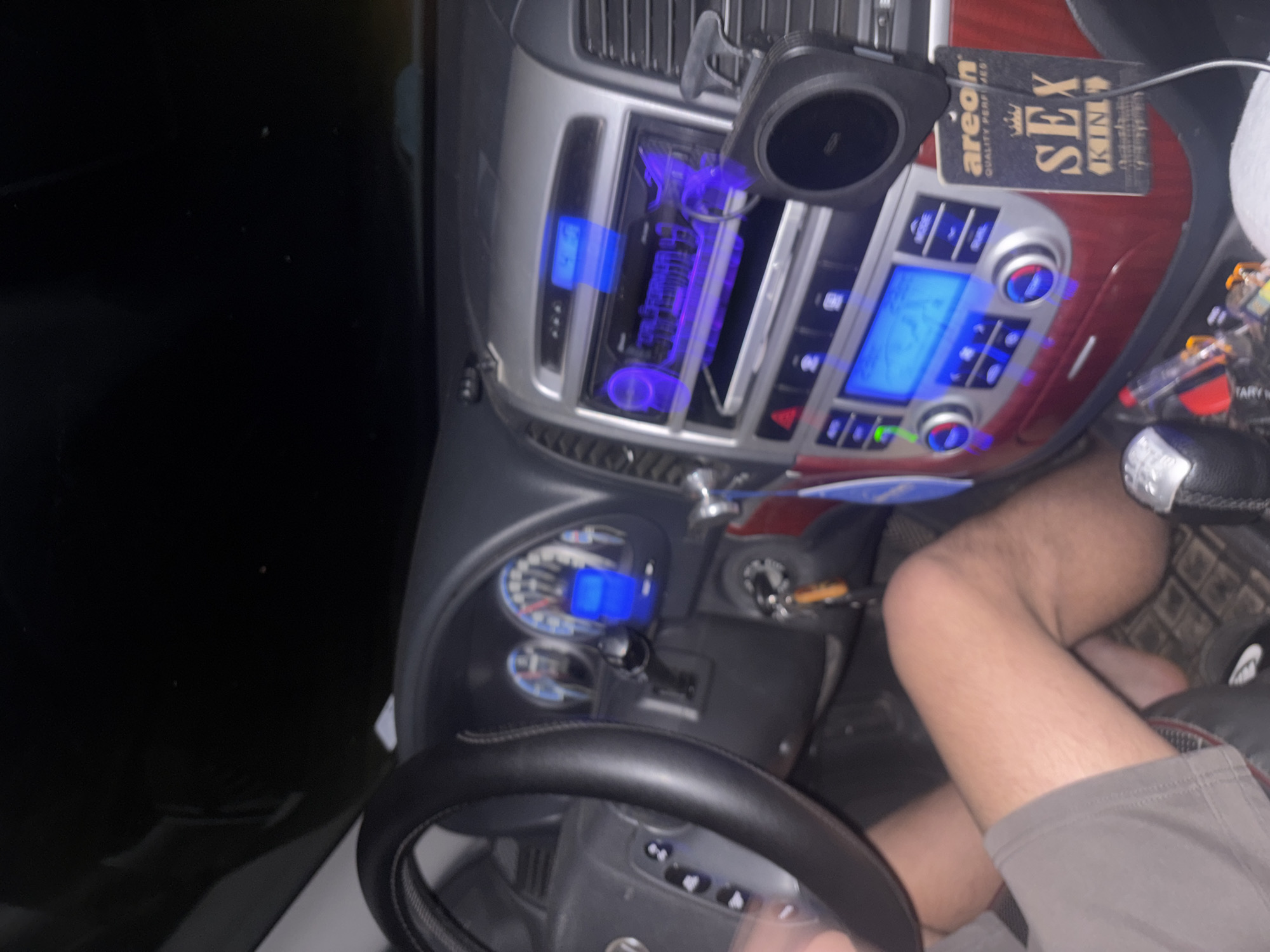 The image depicts the interior of a car at night. The dashboard is visible, featuring blue illuminated displays for the radio and climate control. An air freshener labeled "SEX KING" hangs nearby. The steering wheel is in the foreground. A person's leg, clad in shorts, is also visible. The location, Gromshin, Bulgaria, cannot be verified from the image content.