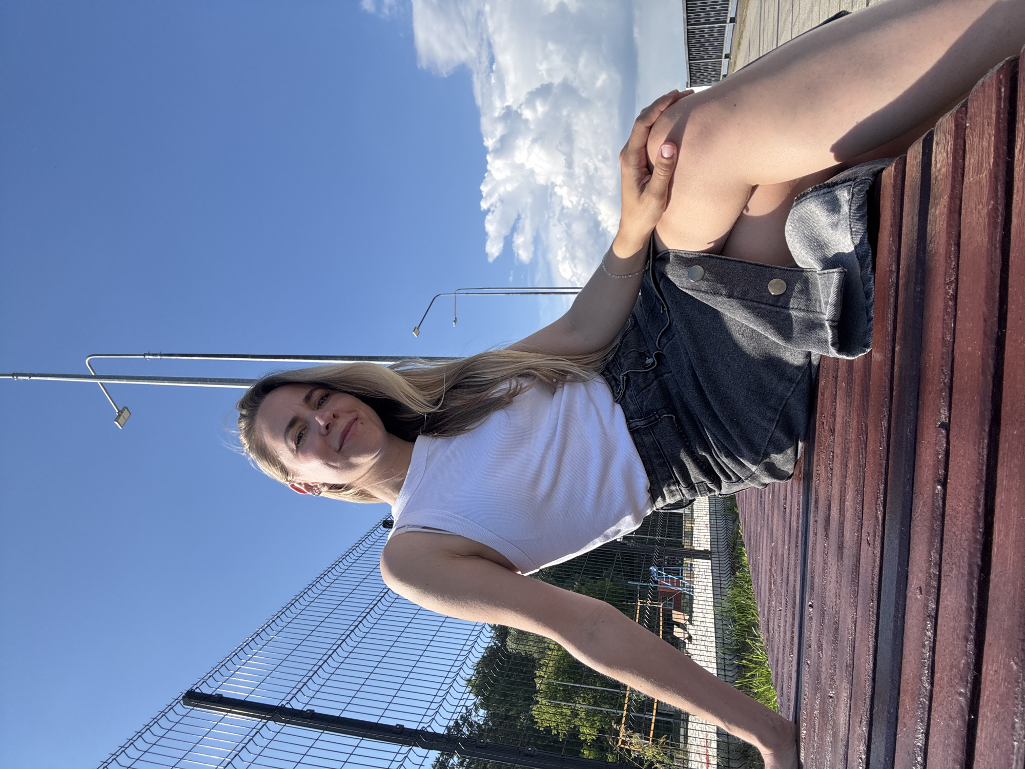 In Chaikovsky, Russia, a young woman with blonde hair and a white tank top sits on a wooden bench. She wears a dark denim skirt and rests her hands on her knees. The bench is located outdoors, near a fenced-off area with playground equipment. In the background, streetlights and a bright blue sky with scattered clouds are visible. The scene is captured in daylight, suggesting a pleasant, sunny day.