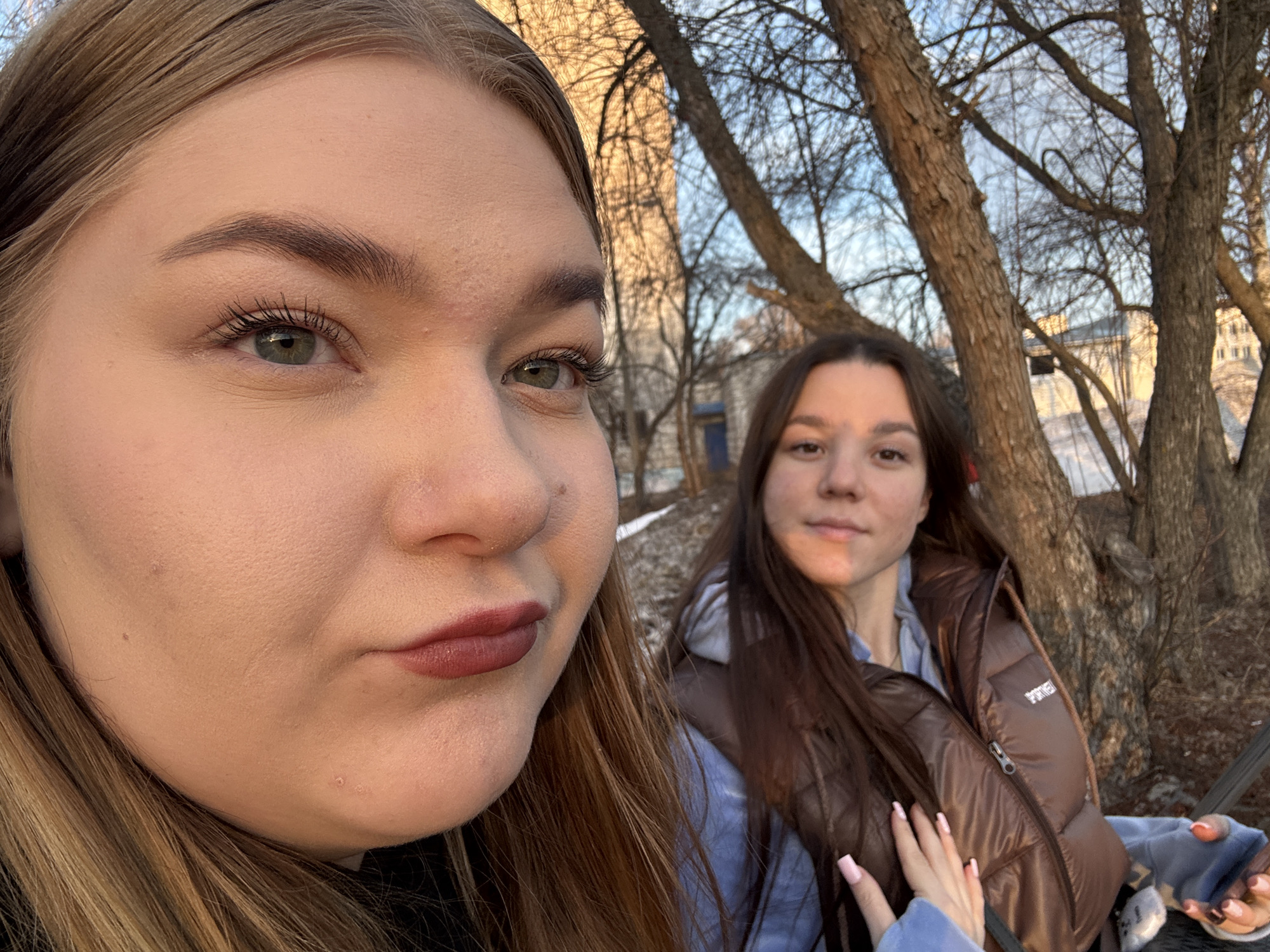 Two young women are photographed outdoors in what appears to be a park or urban green space. The woman on the left is closer to the camera, presenting a close-up view of her face. She has fair skin, light brown hair, and is wearing makeup, including lipstick. The second woman is slightly behind and to the right, with a more distant perspective. She has dark hair and is wearing a brown vest over a light blue shirt. A hand with pink painted fingernails rests on her vest. The background includes trees with bare branches, and a building is faintly visible in the distance.
Without specific landmarks or signs, and with no access to metadata, pinpointing the exact location in Chaikovsky, Russia, is impossible based solely on visual information.