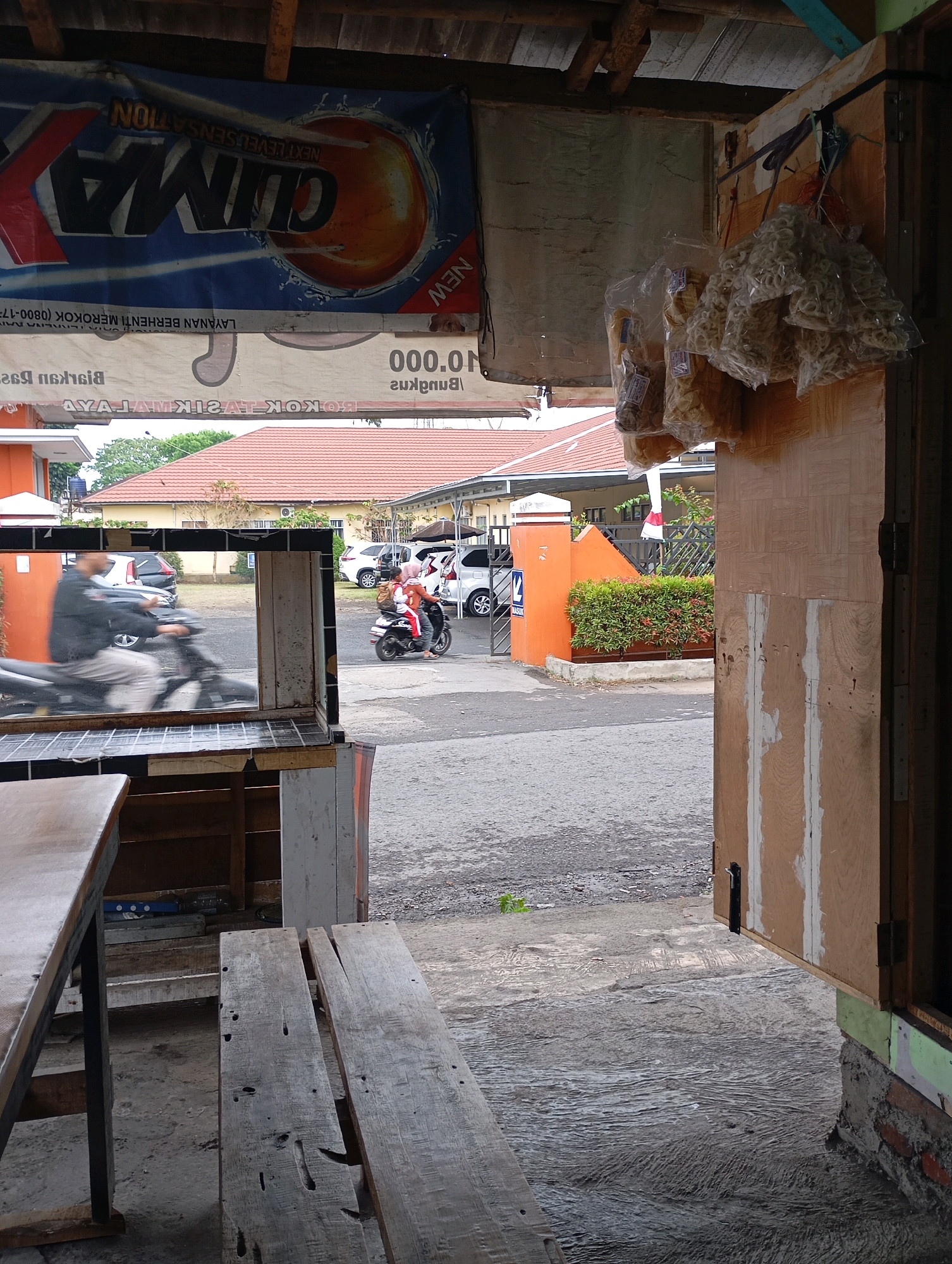 The image depicts an outdoor scene in Tasikmalaya, Indonesia, possibly at a small roadside establishment or warung. In the foreground, a wooden table and bench are visible, suggesting a seating area for customers. To the right, a wooden structure with a partially open door stands, displaying several bags of snacks hanging from it. Beyond this structure, a street can be seen with two individuals on a motorbike. One person is driving, and another is riding as a passenger. A white car is parked on the side of the street, and further in the background, a building with a red tiled roof is visible. The overall environment appears to be a typical urban or semi-urban setting in Indonesia. A banner with the word "CLIMA" is displayed prominently, with Indonesian text suggesting a "Next Level Sensation."