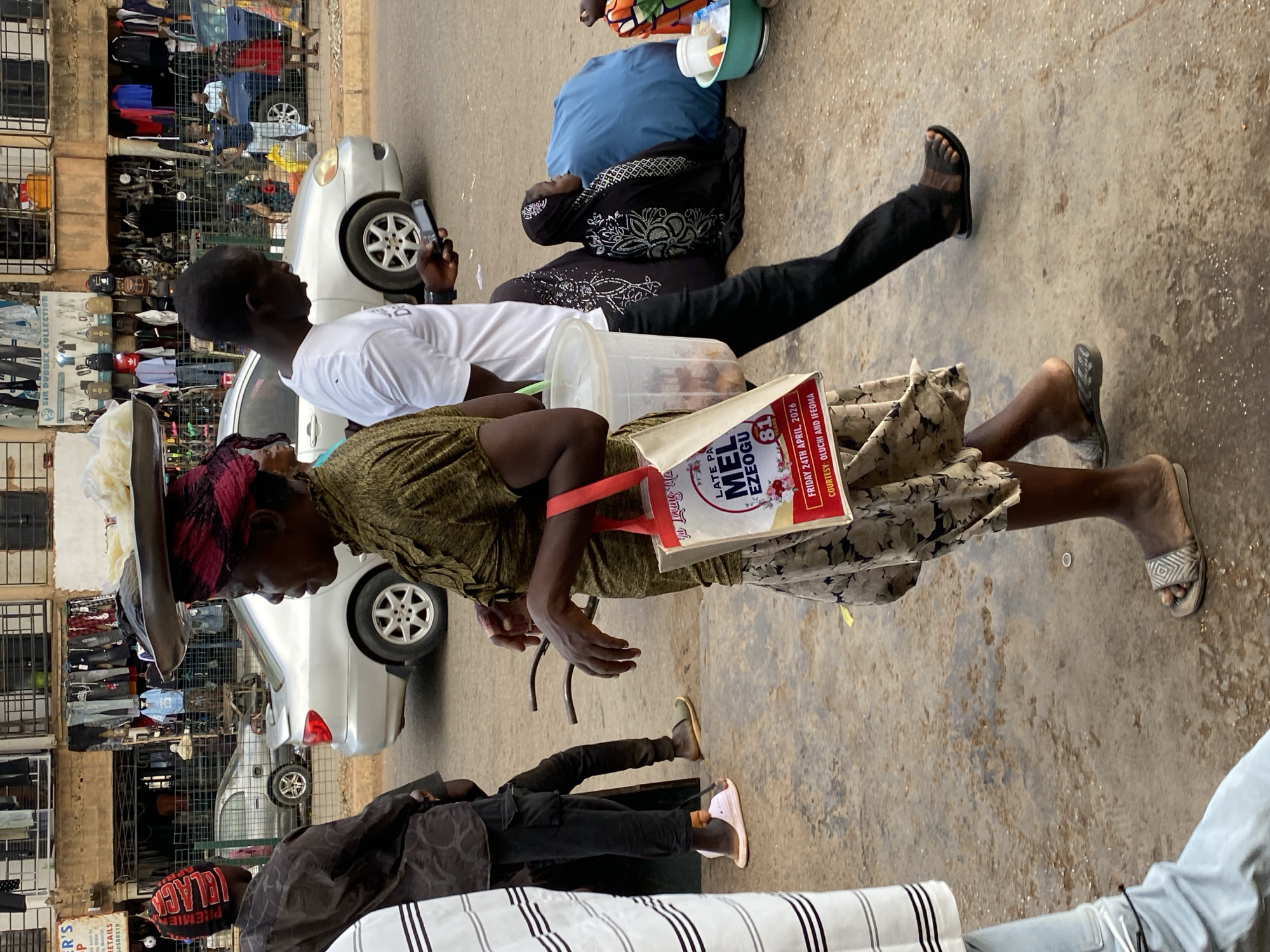 On a city street in Bukuru, Nigeria, several individuals are present. A woman in a patterned dress and sandals carries a shopping bag and wears a hat or head covering. A man next to her wears a white t-shirt and black pants and is looking at a phone. Nearby, a person on the ground is covered with a blue sheet and has a bucket next to them. Another person in black is walking toward the street, wearing sandals. Behind the people are silver cars, and a shop front with clothing displayed in cages. The shopping bag reads "LATE PA MEL EZEOGU". Text on the bag notes "FRIDAY 24TH APRIL, 2026". The ground is a concrete pavement.