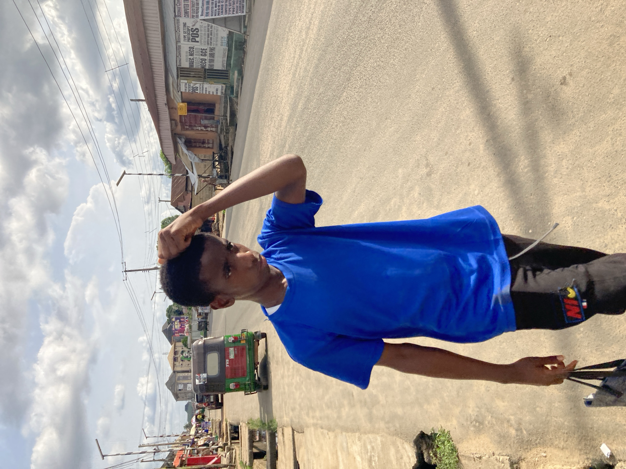 The image features a young man standing on a street in Aba, Nigeria. He is wearing a blue shirt and black pants with a "NI" graphic. He has one hand on his head and the other holds a black strap. Behind him, there is a commercial building with signs for computer services, typing, photocopying, WAEC, NECO, POS, and internet. A green auto-rickshaw with "TVS" displayed is partially visible behind the man. The street appears paved. The sky is partly cloudy. Electrical poles and wires are visible overhead. Further down the street are more buildings and street activity.