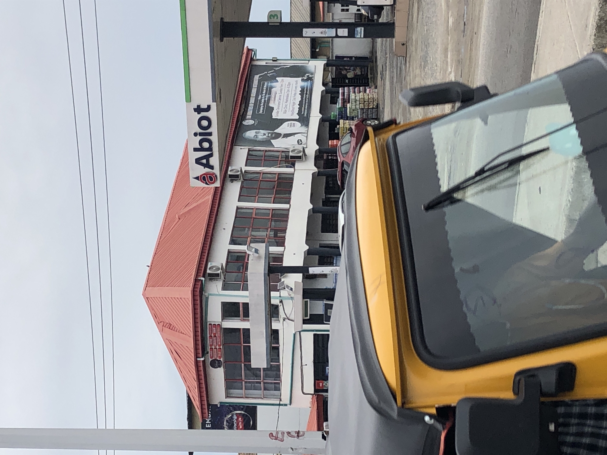 The image captures a partial view of an Abiot filling station in Ado, Nigeria, featuring a multi-story building with a red roof. Visible are the Abiot logo, a large advertisement, windows, and air conditioning units attached to the building's exterior. A yellow vehicle, possibly a tricycle, is prominently in the foreground, obscuring part of the scene. Power lines are also visible against the sky.