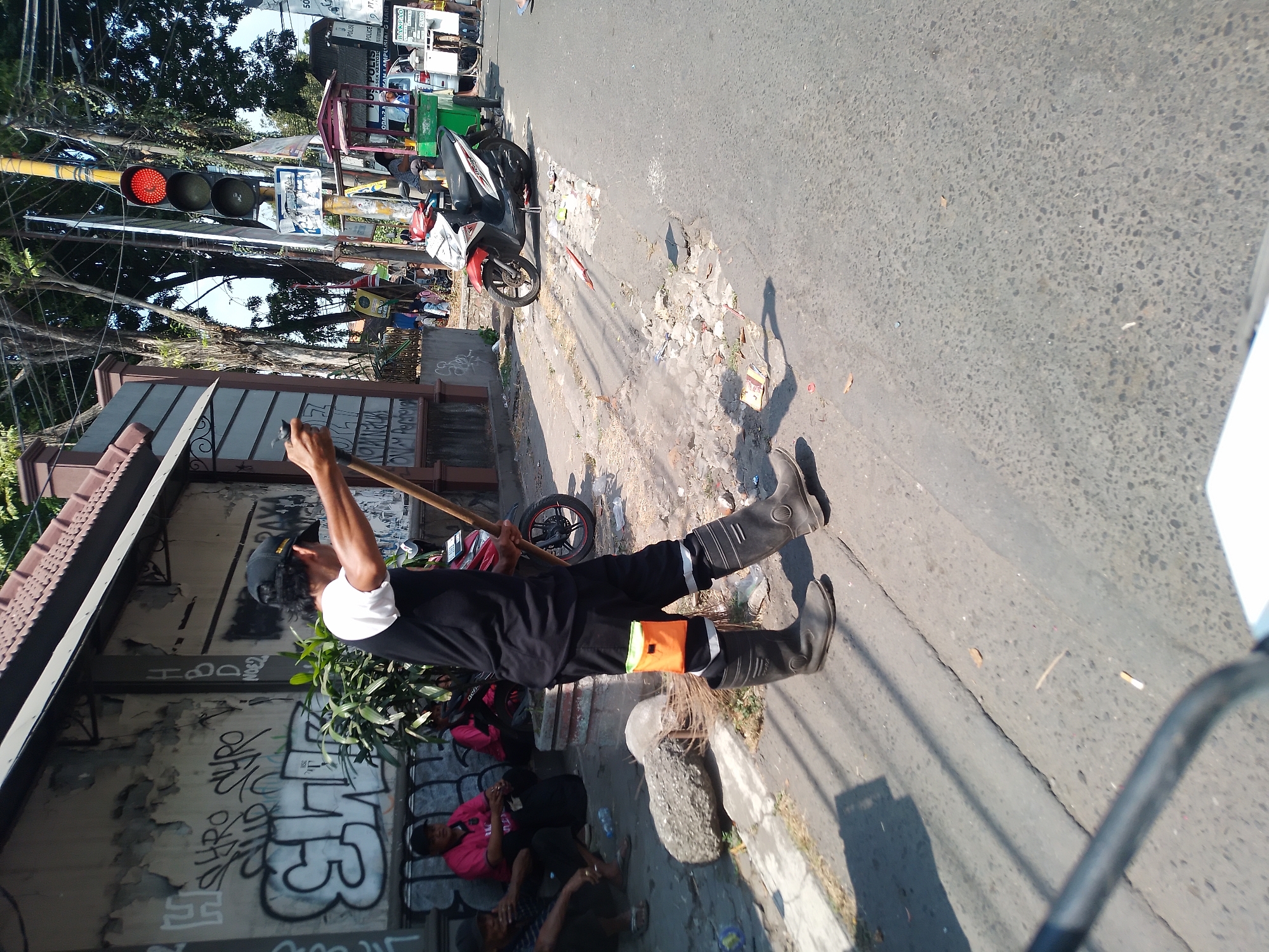 In Kertosono, Indonesia, a street cleaner, wearing a black cap, black vest over a white shirt, black pants with reflective orange and white accents on the legs, and tall black rubber boots, sweeps the street with a broom. He holds a phone in his right hand, seemingly interacting with it while sweeping. The scene takes place on a sunny day, with the street showing signs of wear and tear, including cracks and debris. A red traffic light hangs above, indicating a stop. Several people are visible in the background, some sitting on a low wall and others near a food cart and motorcycles. Graffiti adorns a wall behind the people sitting. The environment suggests a bustling urban setting.