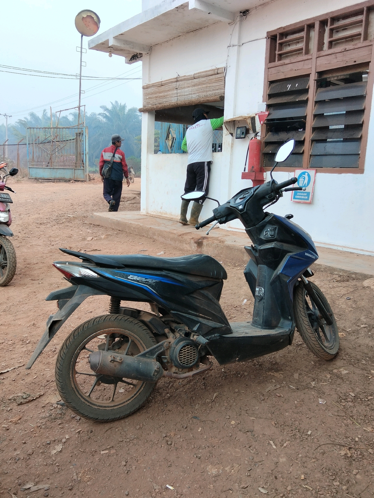 In an outdoor, dusty environment, a blue and black Honda Beat scooter is parked in the foreground, facing right. The scooter has a black seat with blue accents and a silver exhaust pipe. Behind the scooter, a white building with brown-framed louvered windows and a light-colored awning is visible. A red fire extinguisher is mounted on the side of the building near a window. Two individuals are present: one wearing a light green shirt and dark pants is standing at the window of the building, and another person wearing a red and black jacket, dark pants, and carrying a dark bag is walking away from the building towards a blue metal gate in the background. Palm trees are visible beyond the gate, suggesting a tropical or subtropical location. The ground is covered with dirt and scattered debris.
