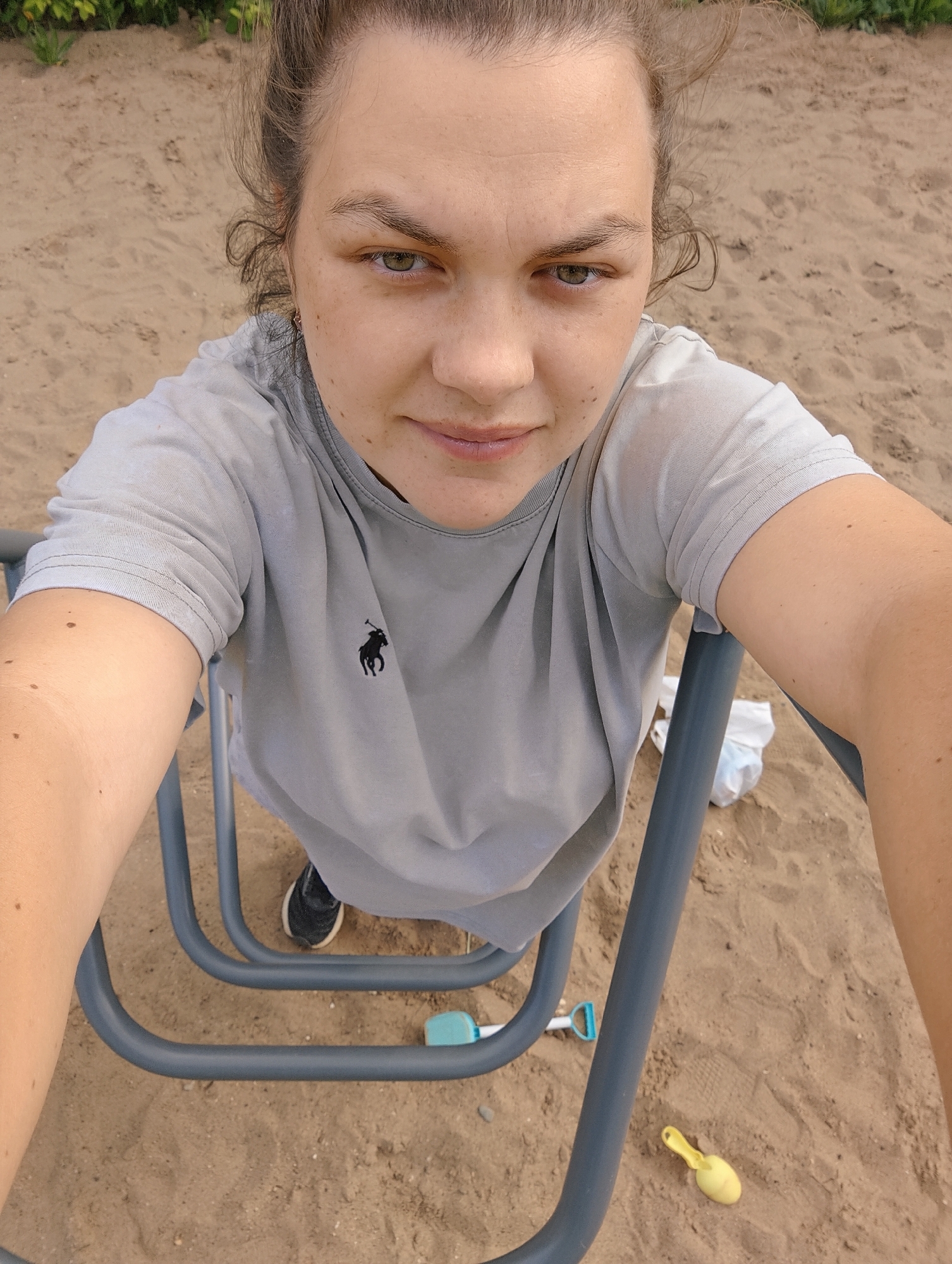 The image is a high-angle, top-down selfie of a person looking directly into the camera. The setting is an outdoor sandy area, resembling a playground or beach, with green foliage visible in the distant background.
The central subject is a person, likely female, with fair skin, dark hair pulled back, and striking green/hazel eyes. Her expression is neutral, direct, and perhaps a little intense or serious as she gazes straight ahead. Visible moles or freckles are scattered across her face and arms. She is wearing a light grey short-sleeved t-shirt, adorned with a small black polo horse logo on the left side of the chest. Her arms are extended, holding onto two parallel, tubular grey metal bars, which appear to be part of a playground structure such as monkey bars or a climbing frame.
Below her, between the metal bars, her feet are visible, clad in black sneakers with white soles resting on the sand. The sandy ground around her shows footprints and appears disturbed. Scattered on the sand are a few items: a small light blue plastic shovel or toy, a bright yellow plastic scoop or shovel, and a crumpled white plastic bag. The bright, natural lighting suggests it is daytime with clear weather. The image captures a casual moment, possibly during recreation in Chaikovsky, Russia.