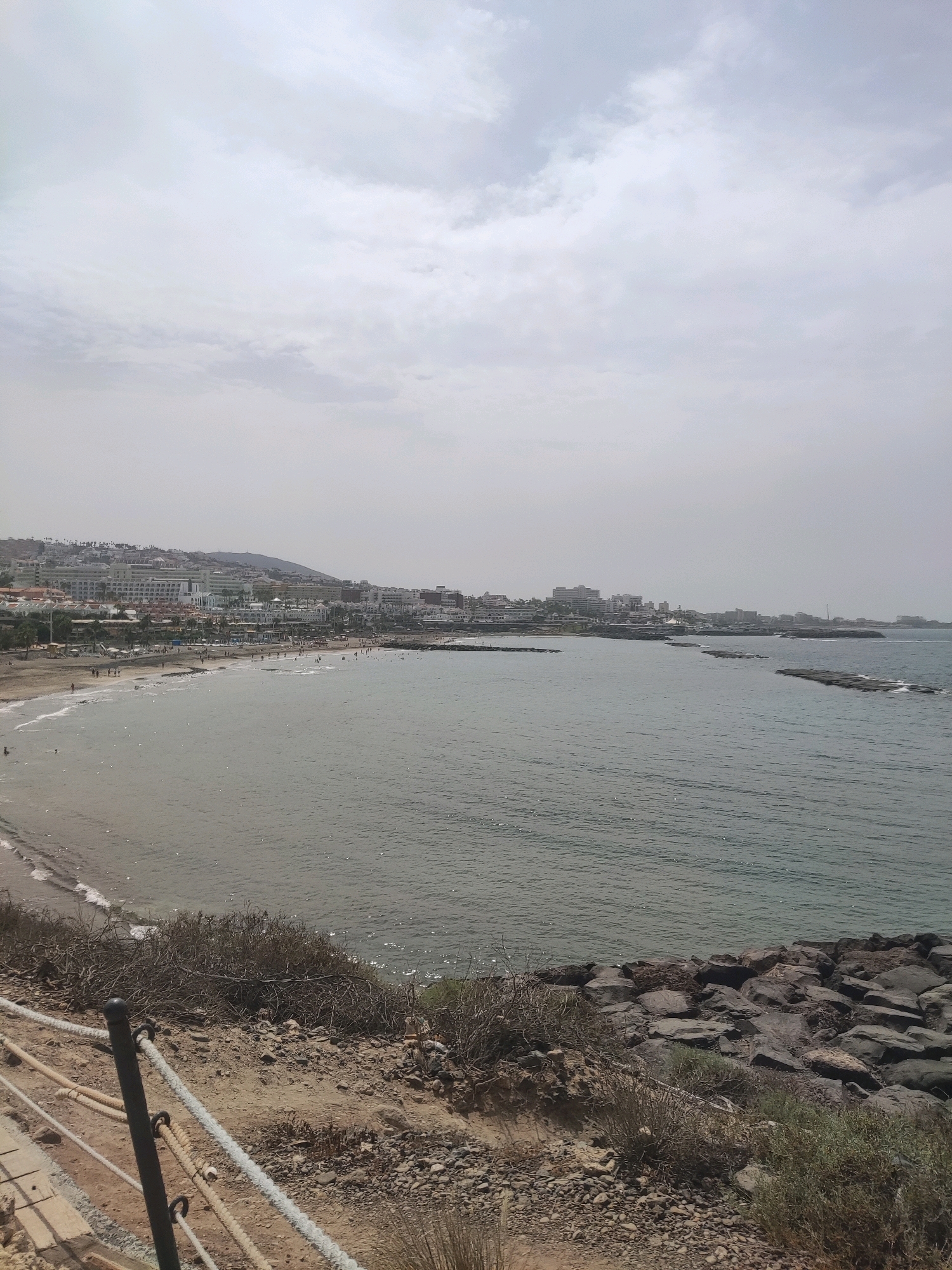 The image shows a panoramic view of a beach and coastline in Costa Adeje-playas Del Duque, Spain. The foreground features a rocky embankment with sparse vegetation and a rope barrier. The beach, populated with numerous individuals, stretches along the coast, with the gentle waves of the sea lapping at its shore. In the background, a dense collection of buildings, likely hotels and resorts, forms the city skyline. The sea is calm, with a few rocky breakwaters extending into the water. The sky is overcast with soft, diffused light.