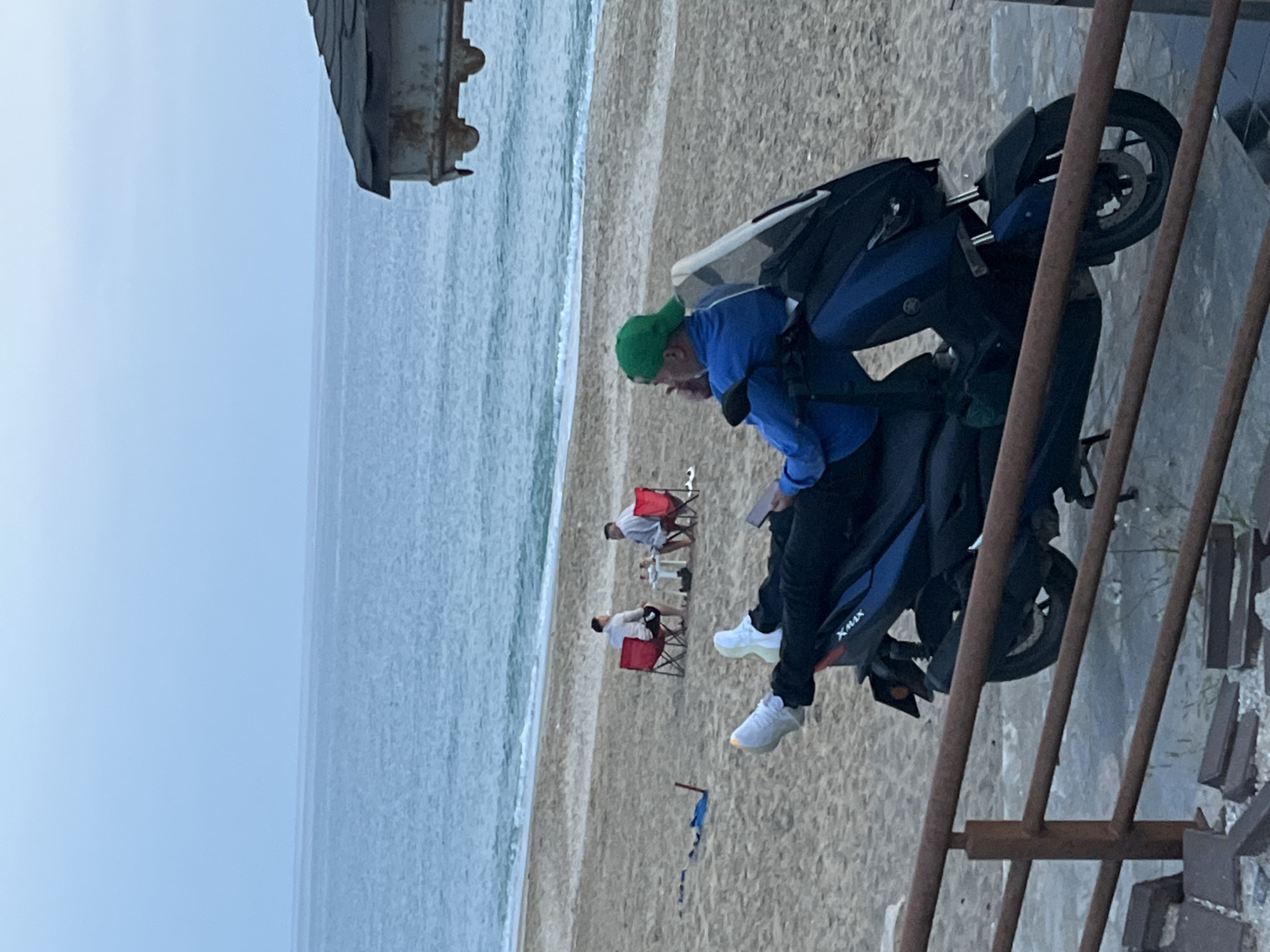 Captured in Alanya, Turkey, the scene unfolds on a beach, where a person wearing a green cap, blue jacket, and black pants is seated on a blue and black Yamaha XMAX scooter. Two people are relaxing in red beach chairs in the distance. Near the scooter, a rusted metal structure is visible, adding a sense of age to the scene. The backdrop reveals a calm ocean under an overcast sky. A small piece of cloth is stuck in the sand.