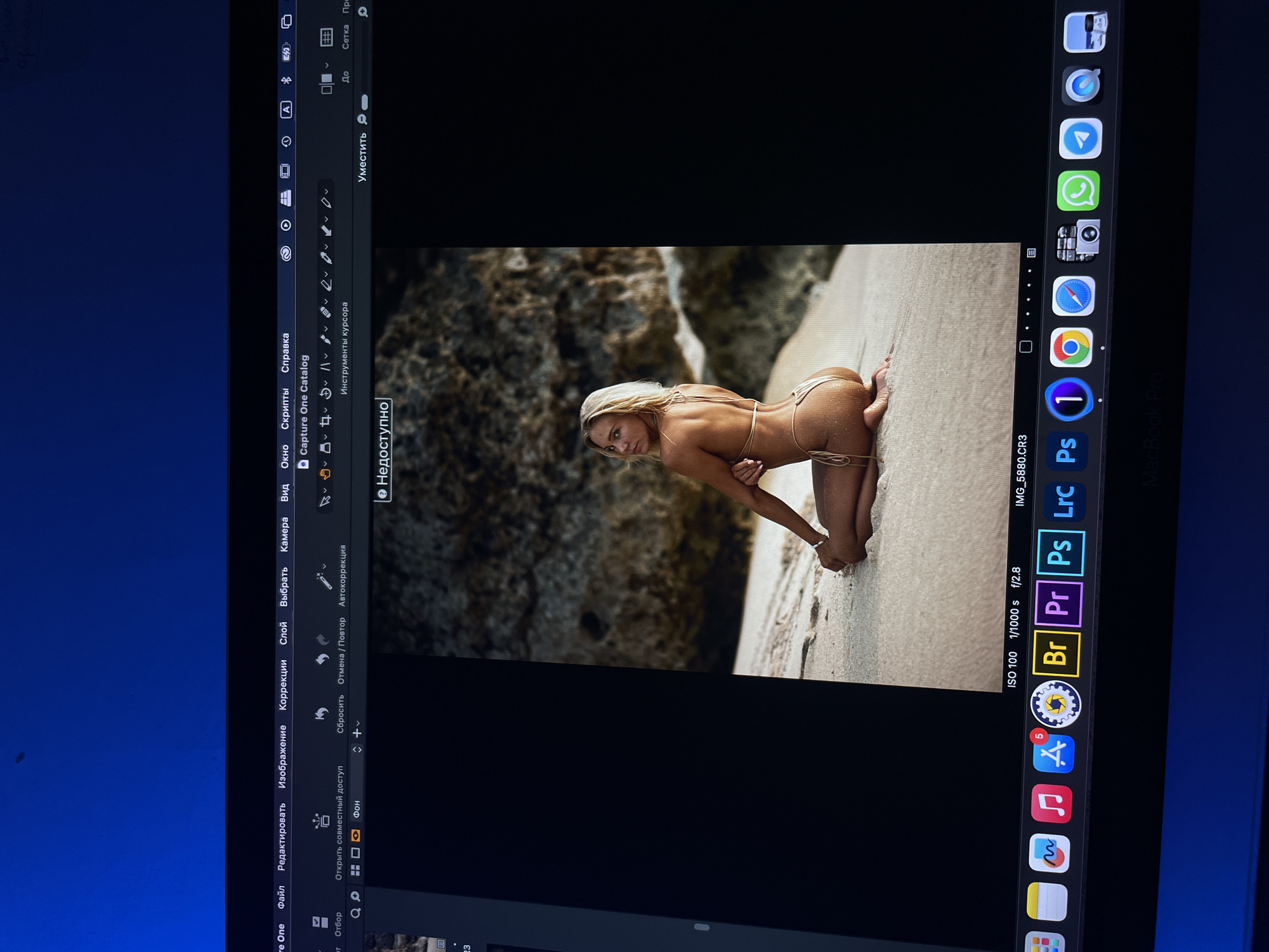 The image shows a woman on a beach, kneeling on the sand. She is wearing a small bikini. Behind her, there is a rocky cliff face. The image is displayed on a computer screen running the Capture One photo editing software on a Macbook Pro. There are several Adobe Creative Suite apps visible on the Mac OS dock, including Lightroom, Photoshop, Premiere, and Bridge. The file name is "IMG_5880.CR3" and the camera settings are listed as ISO 100, 1/1000 second shutter speed, and f/2.8 aperture. Kazan, Russia is not identifiable in this image.